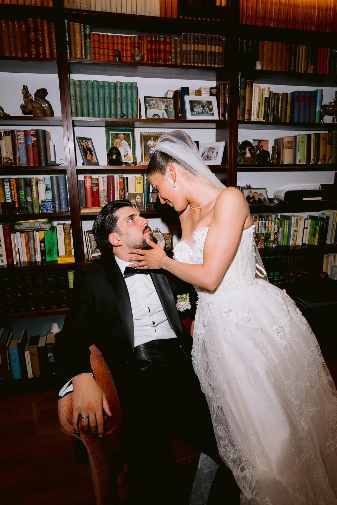 Bride in white lace gown tenderly touches groom's face as he sits in chair before a wood bookshelf library backdrop.