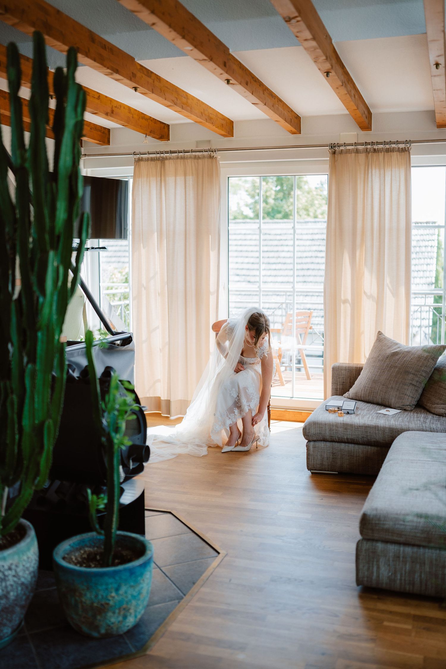 Bride in white wedding gown being helped with dress in bright modern living room with wooden ceiling beams.