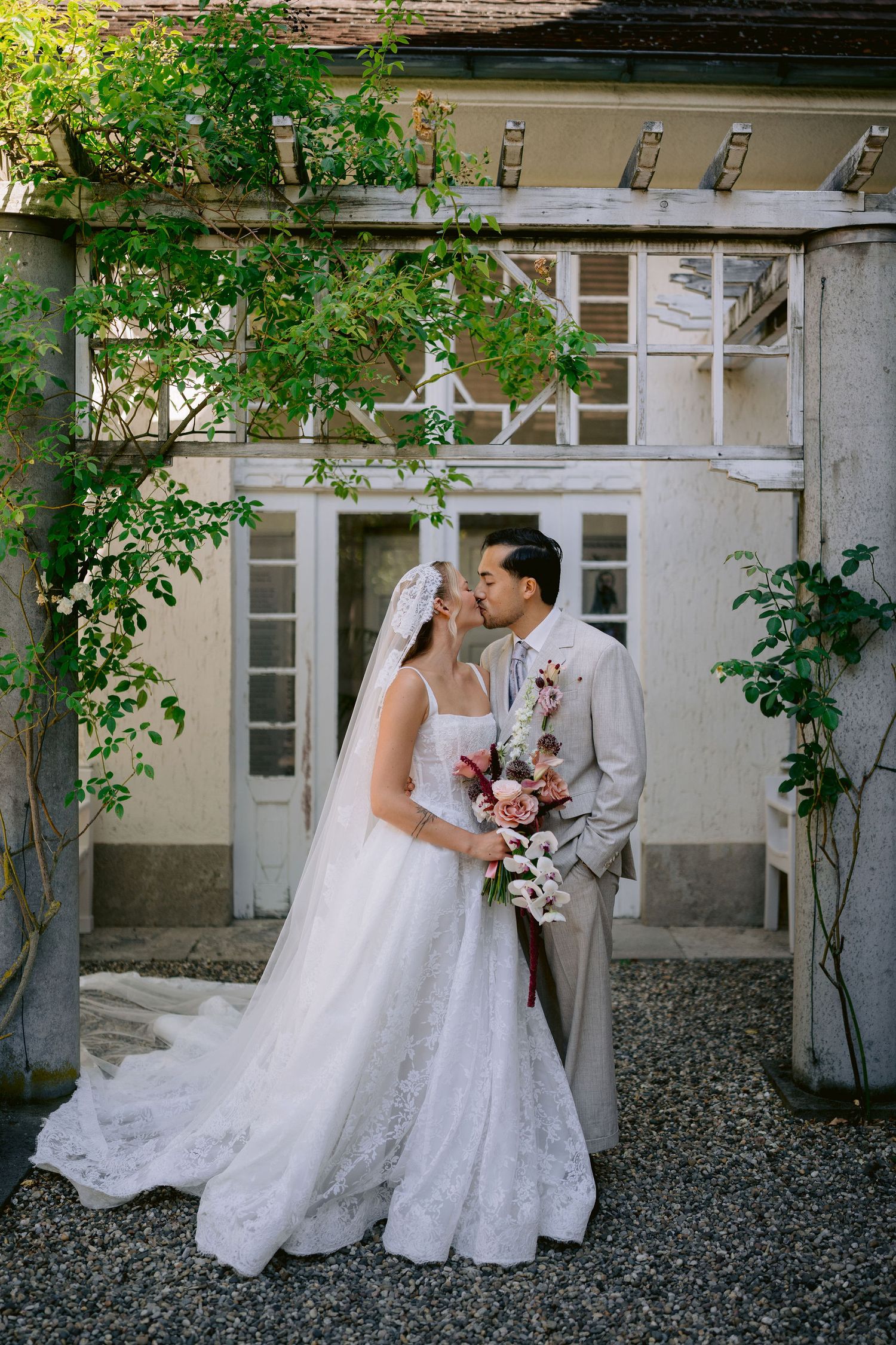 Bride and groom share a kiss outdoors, surrounded by lush greenery, as bride holds a pink and white bouquet.