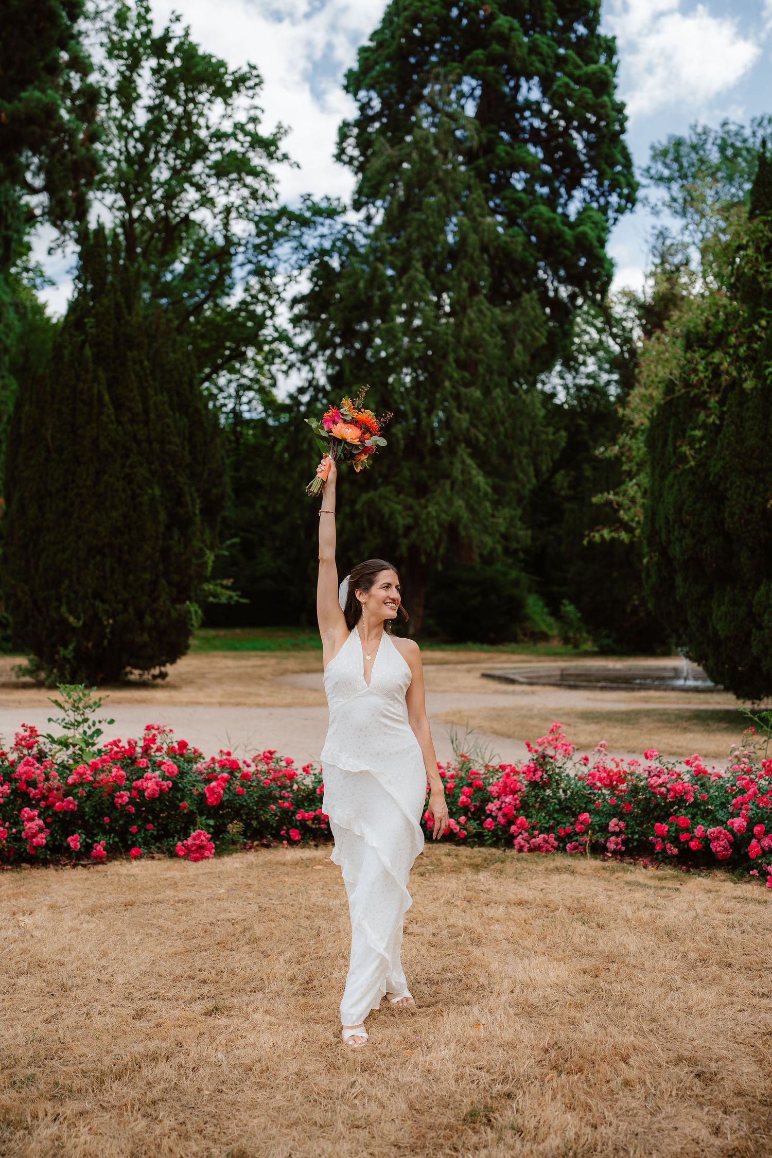 Joyful bride in white halter gown raising colorful bouquet in garden with pink roses and tall green trees.
