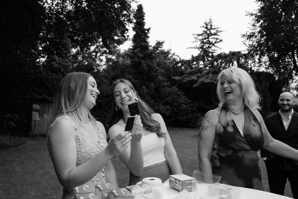 Three people share a joyful moment while drinking and laughing together outdoors at a black and white garden party.