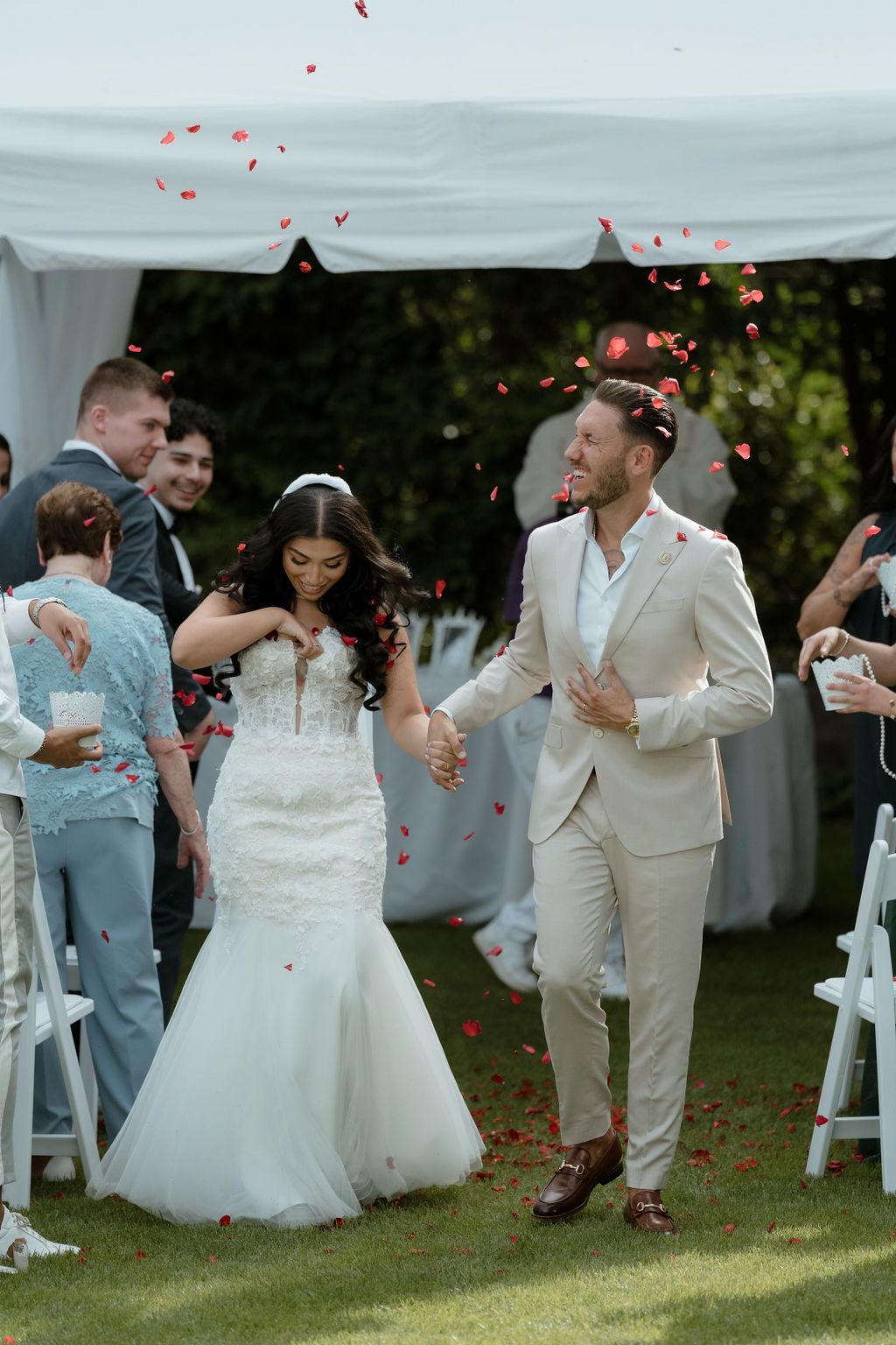 A newlywed couple walks down the aisle under red rose petals while guests celebrate at an outdoor garden wedding ceremony.