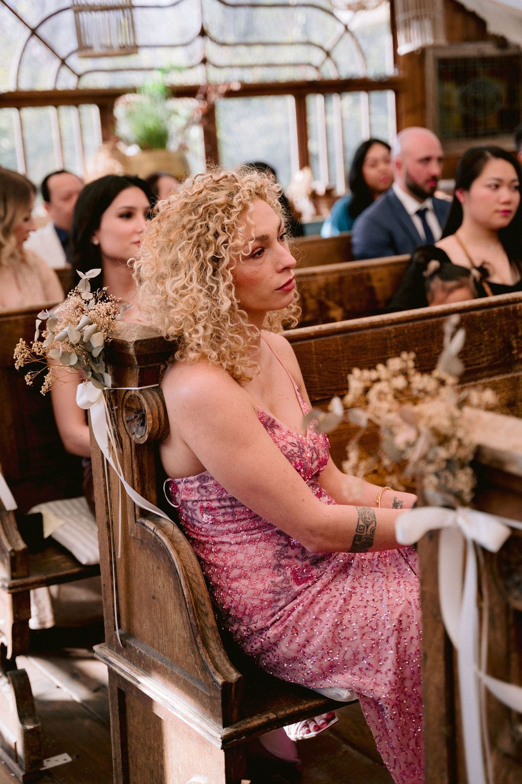 Wedding guests sit in rustic wooden pews decorated with floral arrangements inside a vintage venue.