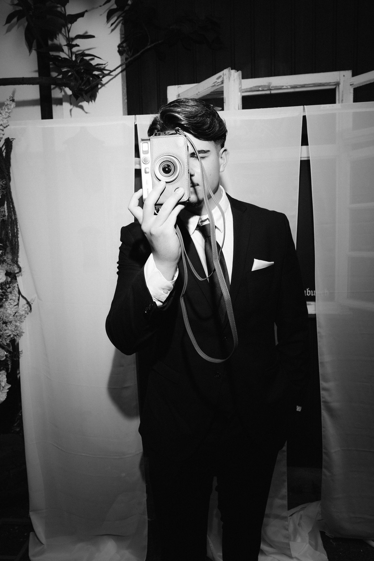 Black and white photo of a young man in a suit holding a vintage camera up to his face at a formal event.