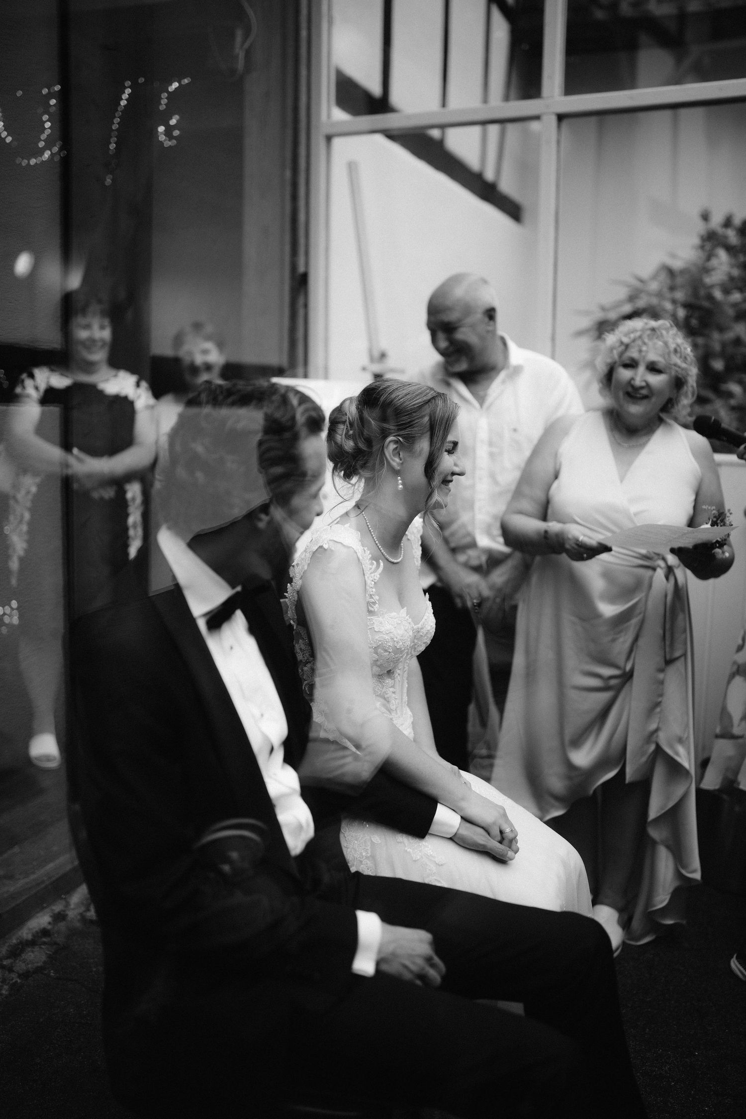 Bride sits on grooms lap laughing during wedding reception as guests look on in black and white photo.