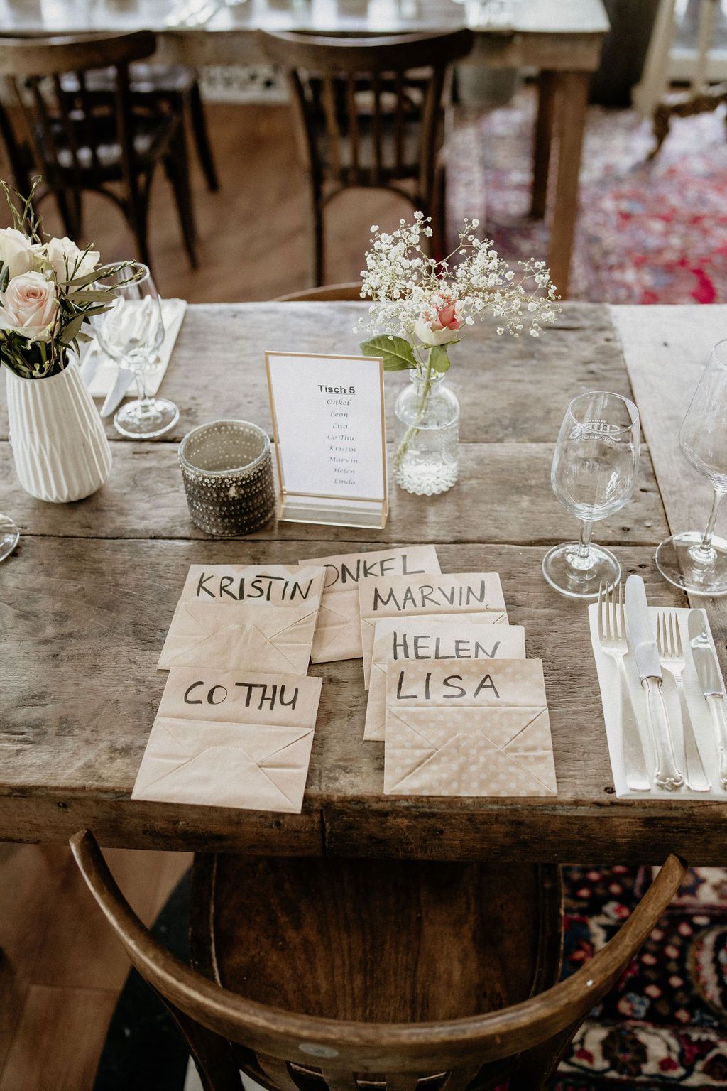 Place cards with handwritten guest names on rustic wooden table settings with delicate flowers in glass vases at a wedding.
