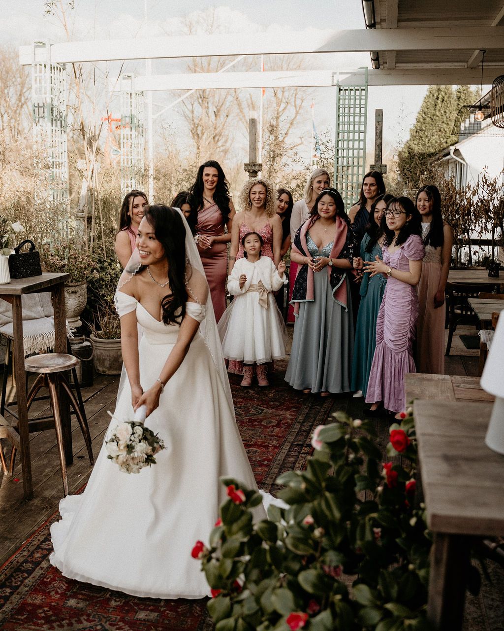 A wedding party gathers on a porch decorated with vintage rugs and greenery as they pose together in pastel dresses.