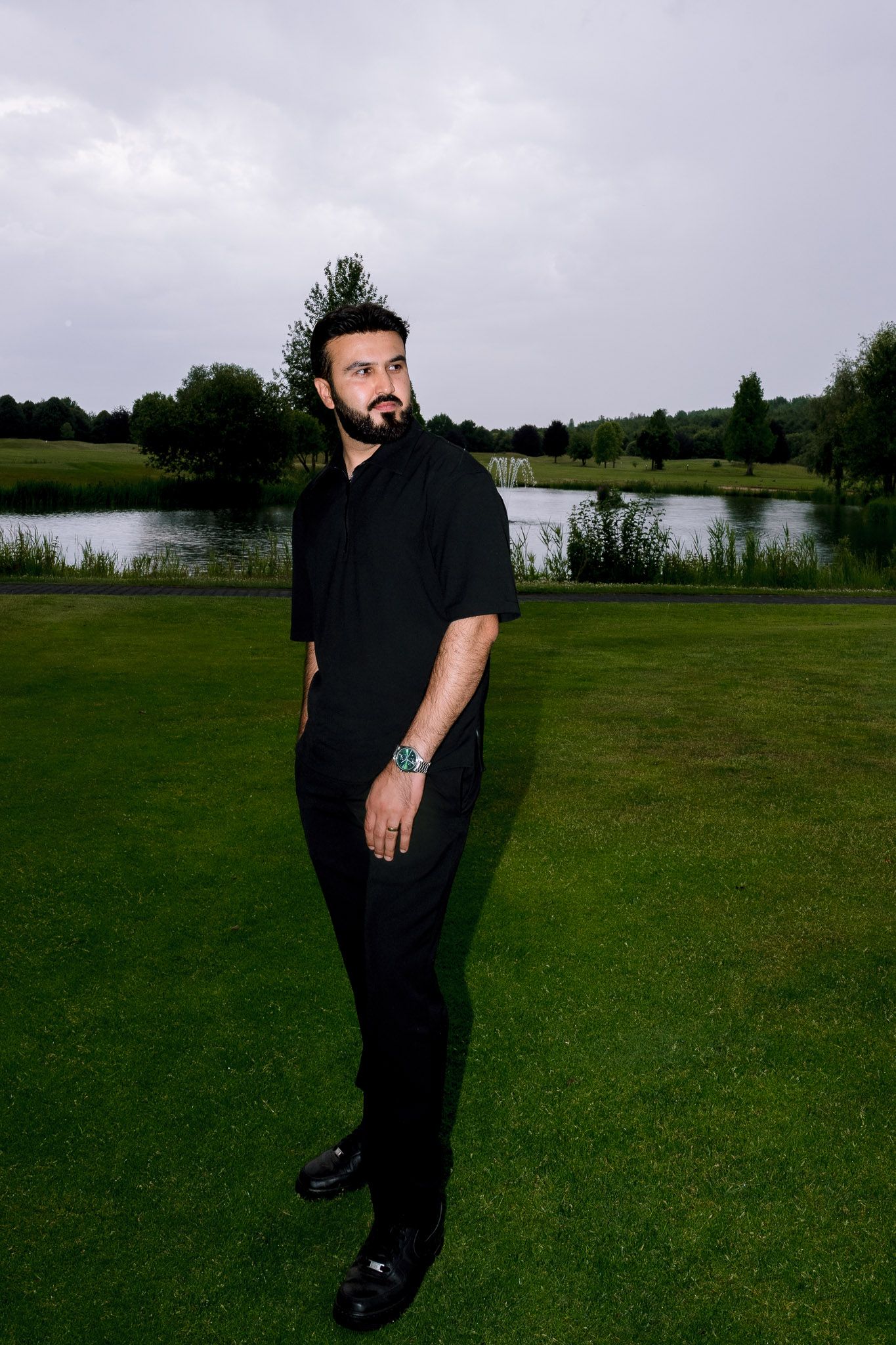 Man in black outfit standing on lush green grass near a pond with trees under overcast sky.