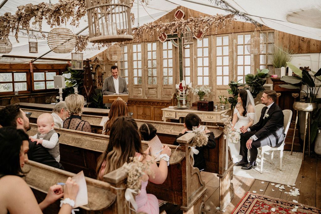 People gather in a vintage greenhouse-style venue with antique furniture and warm lighting for a social event.