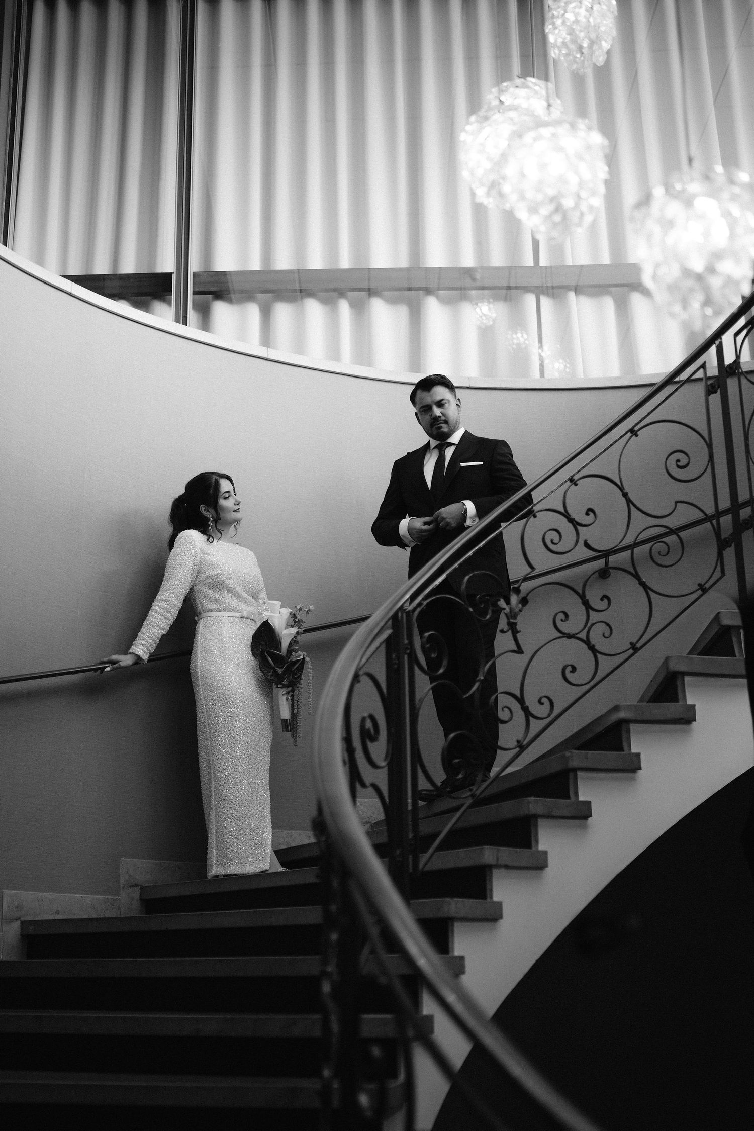 Elegant black and white wedding photo of bride and groom on ornate staircase with crystal chandeliers.