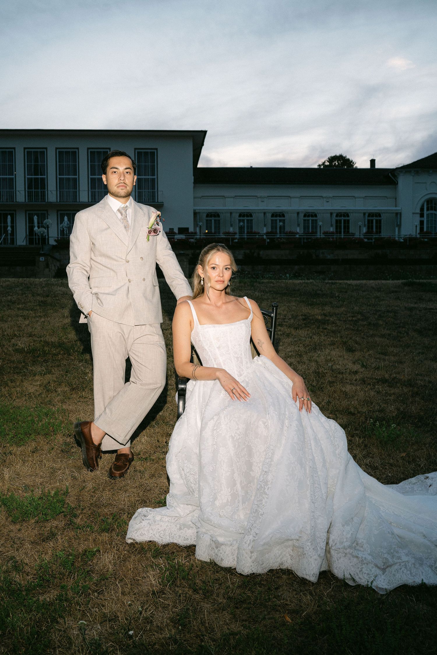 Bride seated in white ballgown and groom standing in beige suit pose before white wedding venue at dusk.