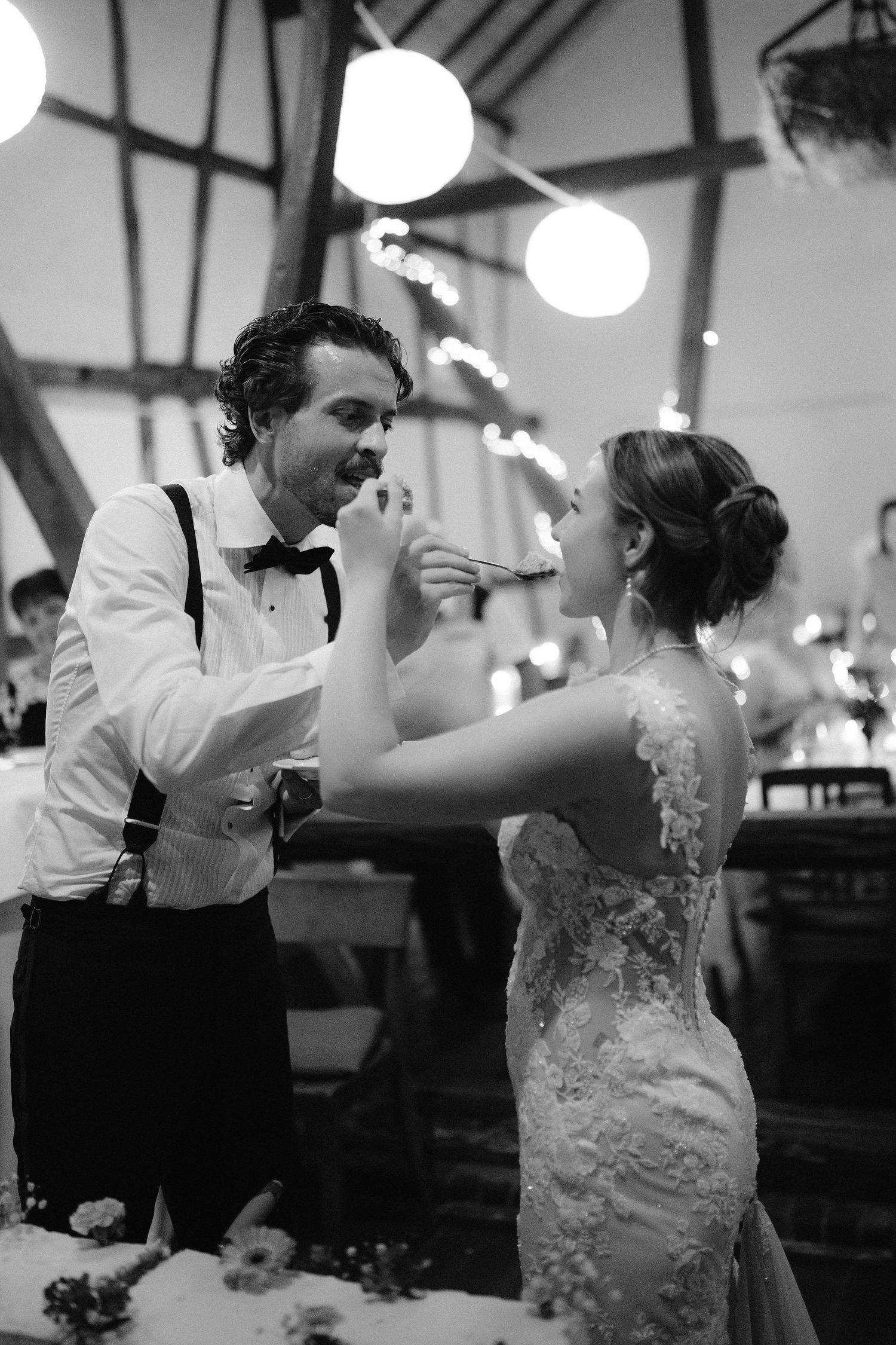 Black and white photo of bride and groom feeding each other wedding cake at elegant barn reception.