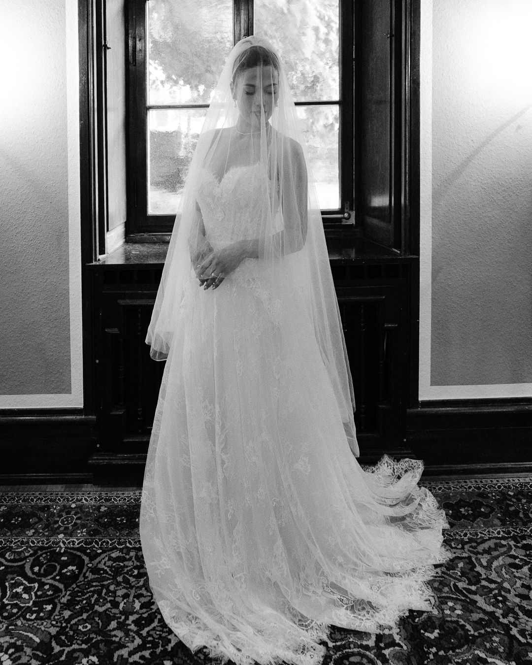 Elegant bride in strapless lace gown and flowing cathedral veil stands before a window in a classic black and white photo.