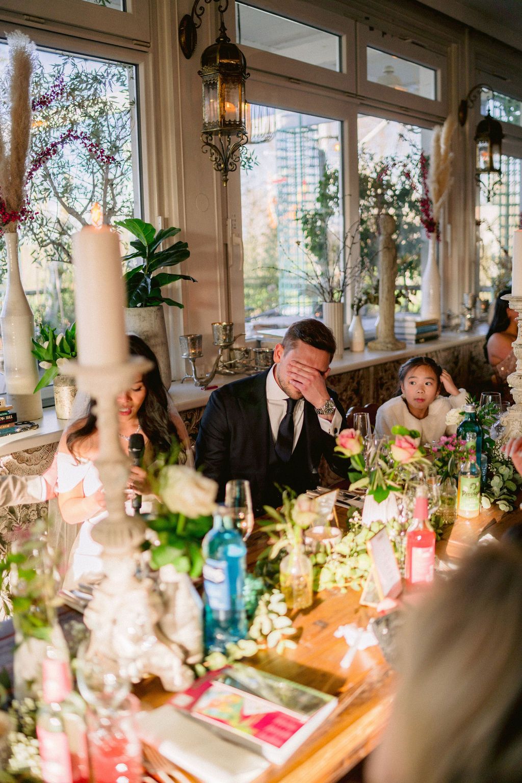 Natural light streams through windows at a joyful wedding reception dinner with decorative table settings and glowing lanterns.