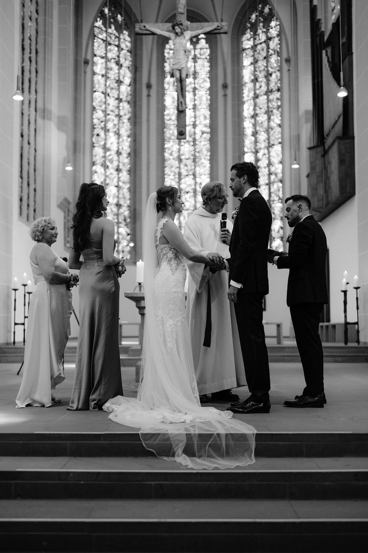 Black and white wedding ceremony photo in a church with stained glass windows, bride and groom exchanging vows at the altar.