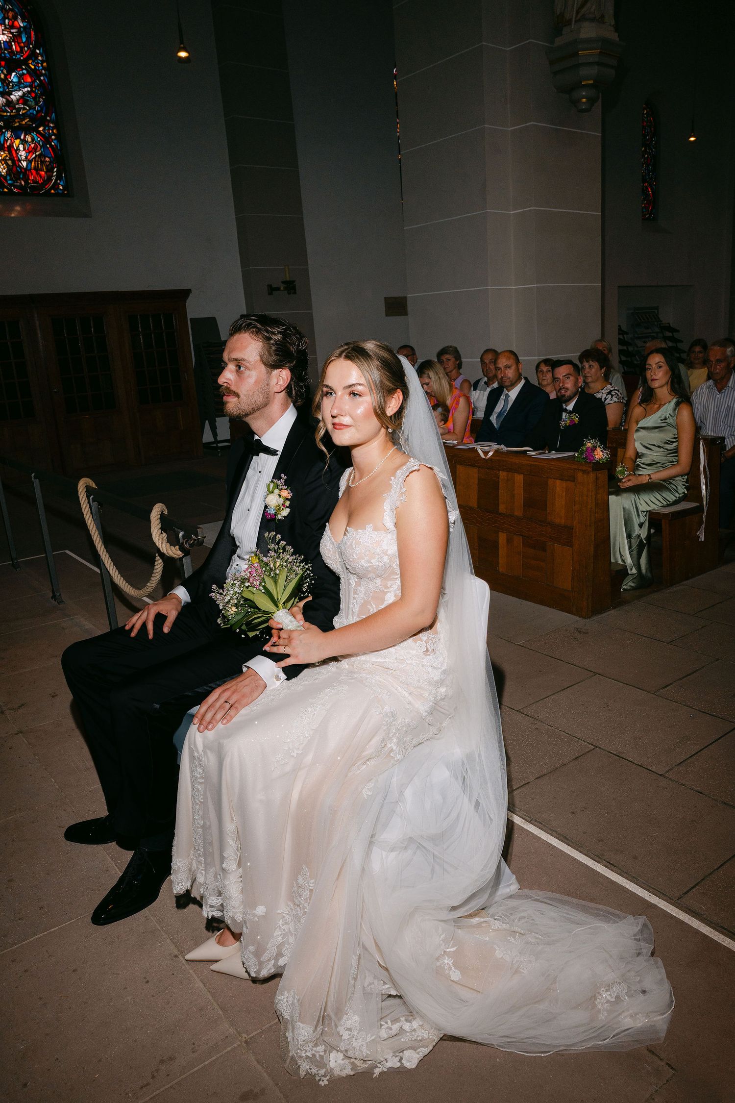 Bride and groom seated at church altar during wedding ceremony, bride in lace gown holding bouquet.