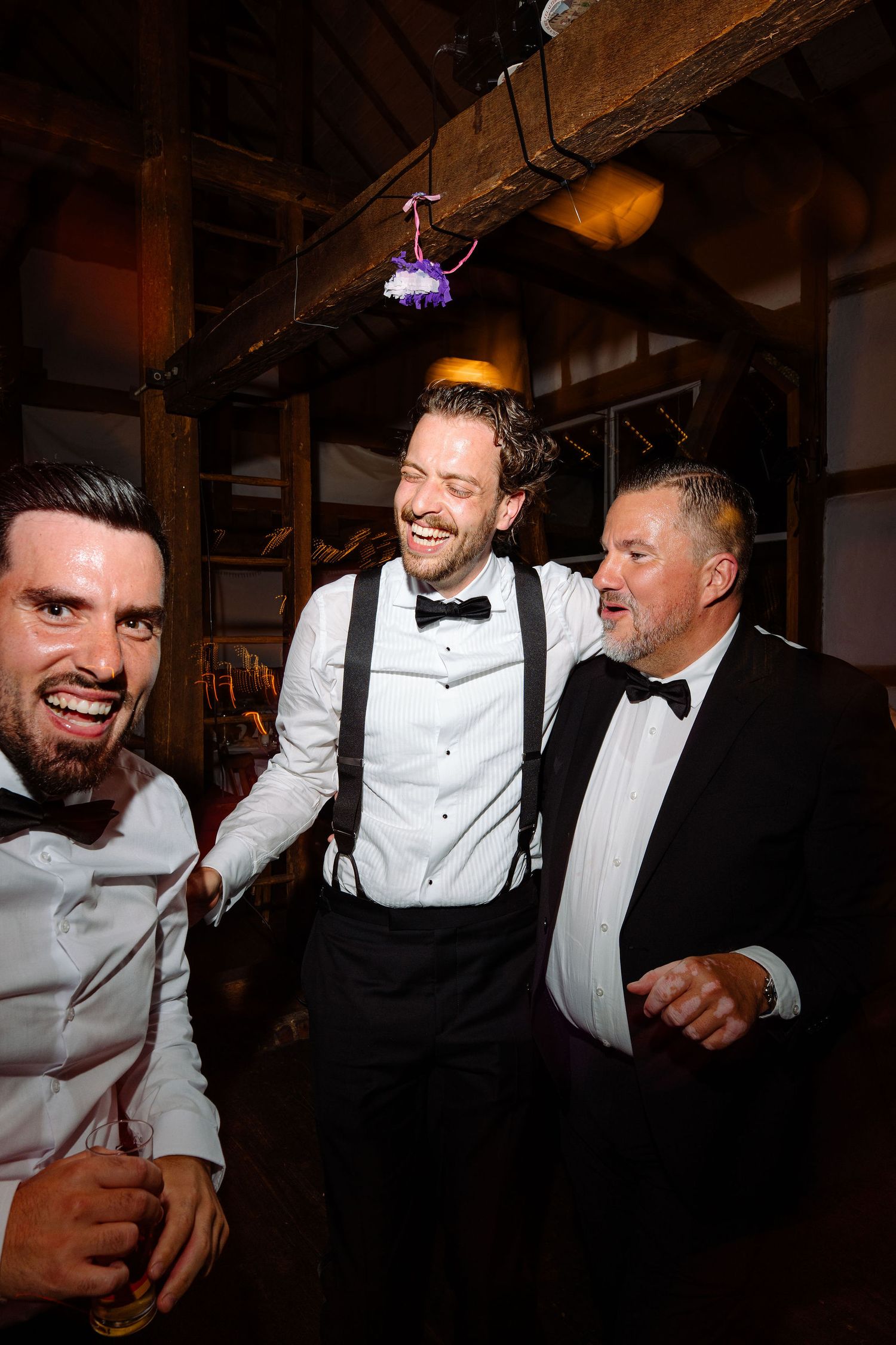 Three men in formal attire laughing and celebrating together at a wedding reception in a rustic venue.