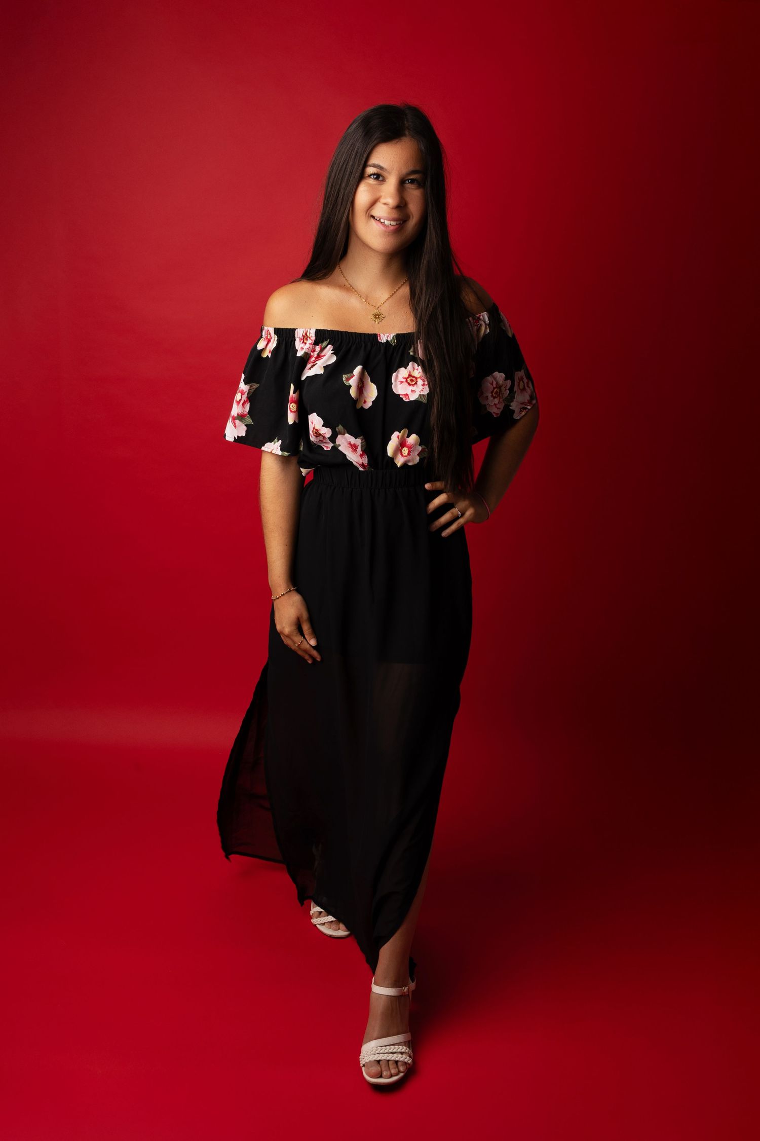 A person wearing a black off-shoulder dress with pink floral details poses against a deep red studio backdrop.