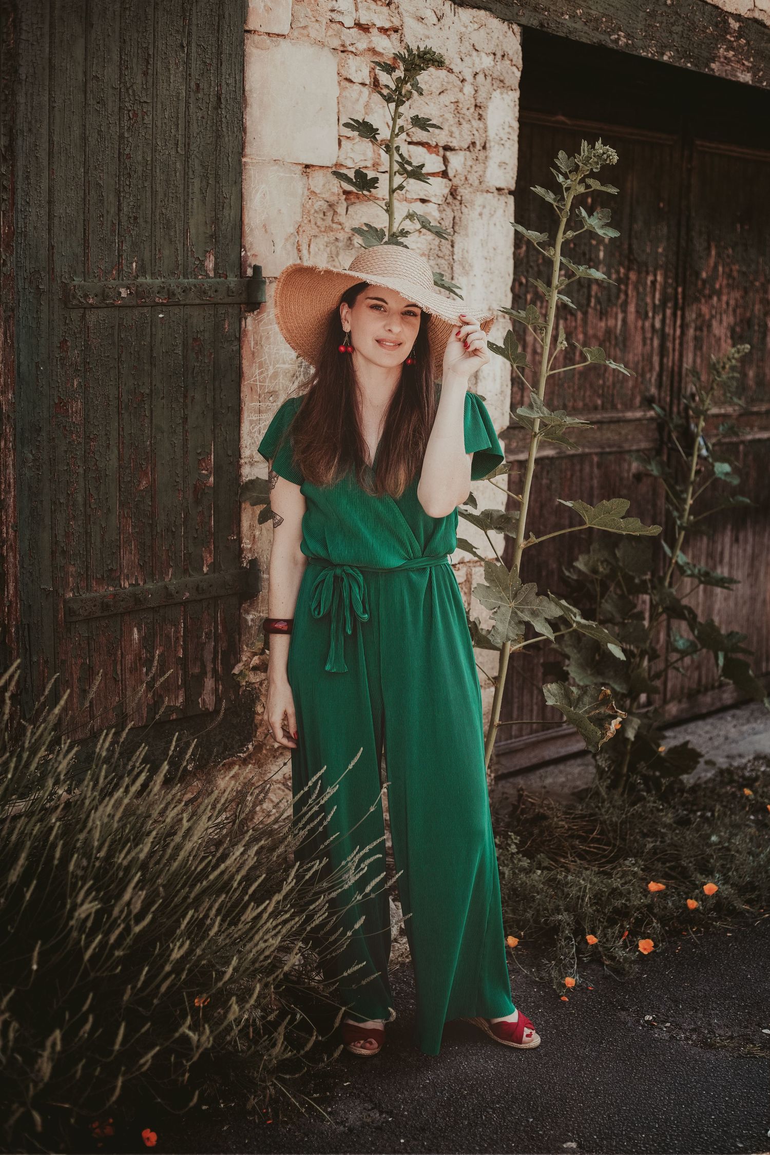 Woman in green jumpsuit and wide-brim hat posing outdoors near rustic wooden doors and stone wall.