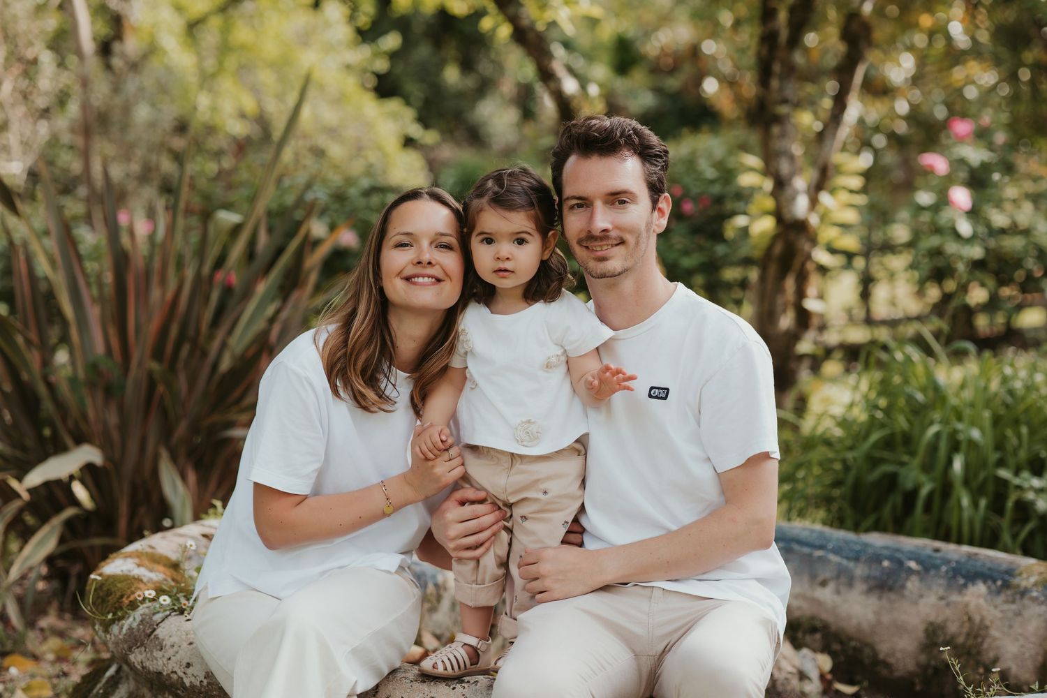 Happy family of three posing outdoors in white outfits, parents holding toddler girl in lush green garden setting.