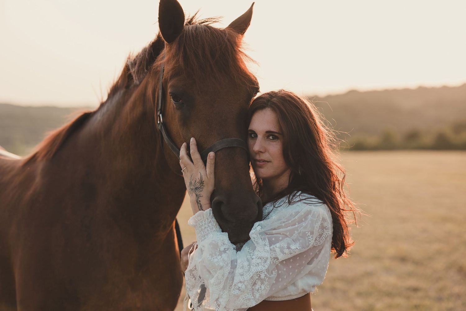 Woman in white lace dress stands in golden field with horse at sunset creating romantic silhouette.
