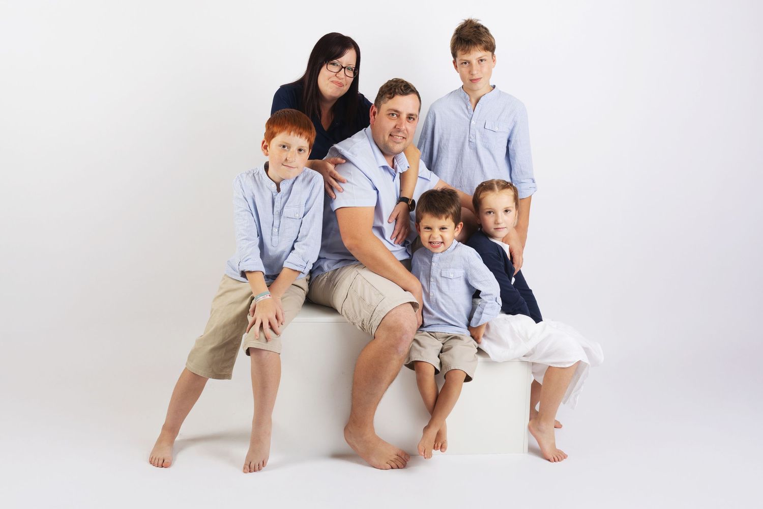 A family group sits together on a white background wearing coordinated light blue and white casual clothing.