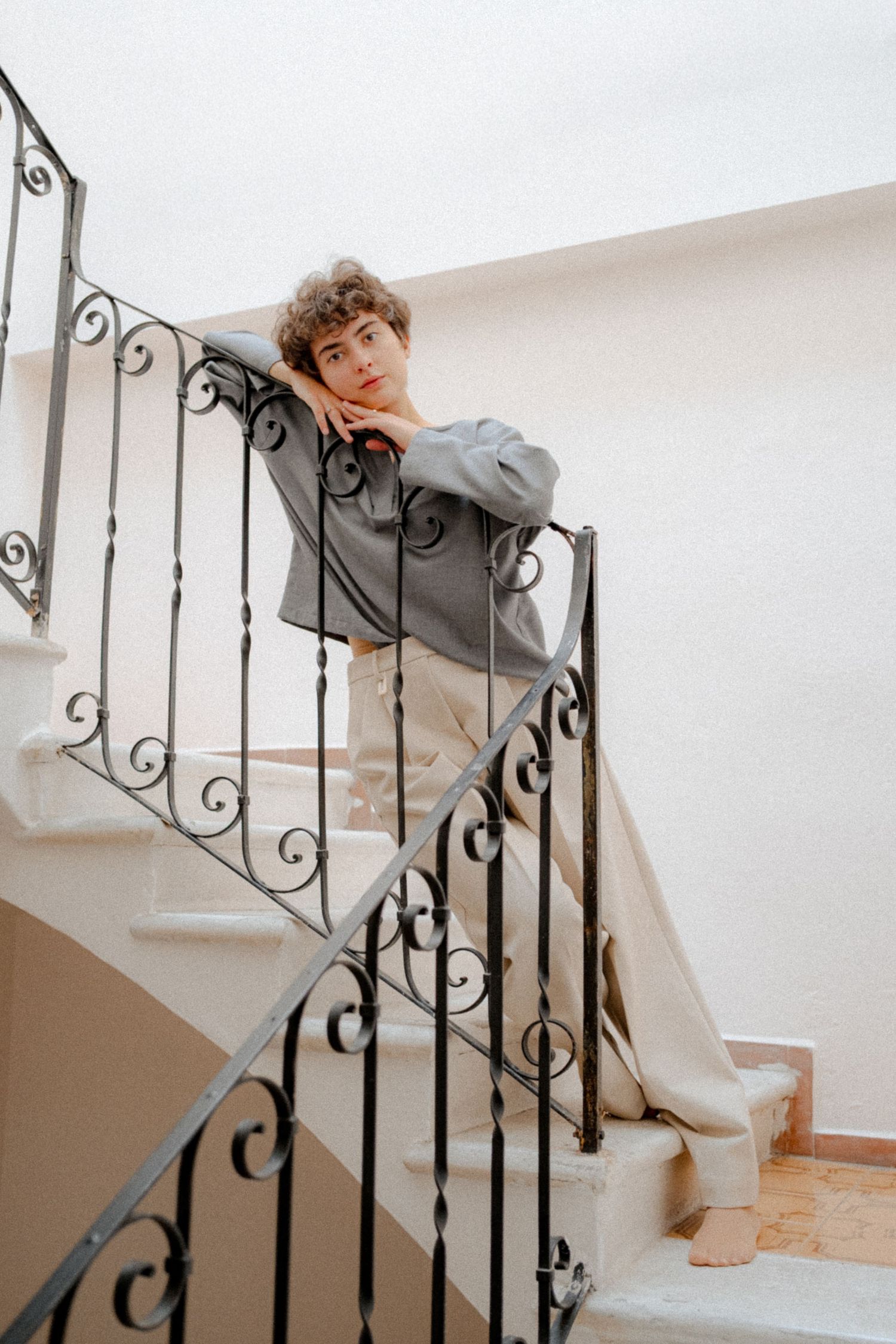 Someone in a gray sweater and beige pants poses on an elegant spiral staircase with ornate wrought iron railings.