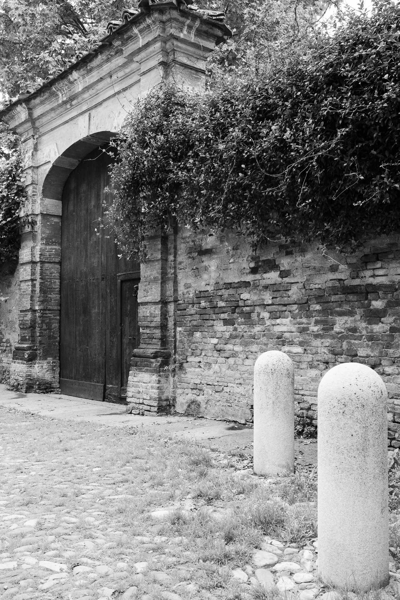 Stone archway entrance with brick wall and concrete bollards in black and white.