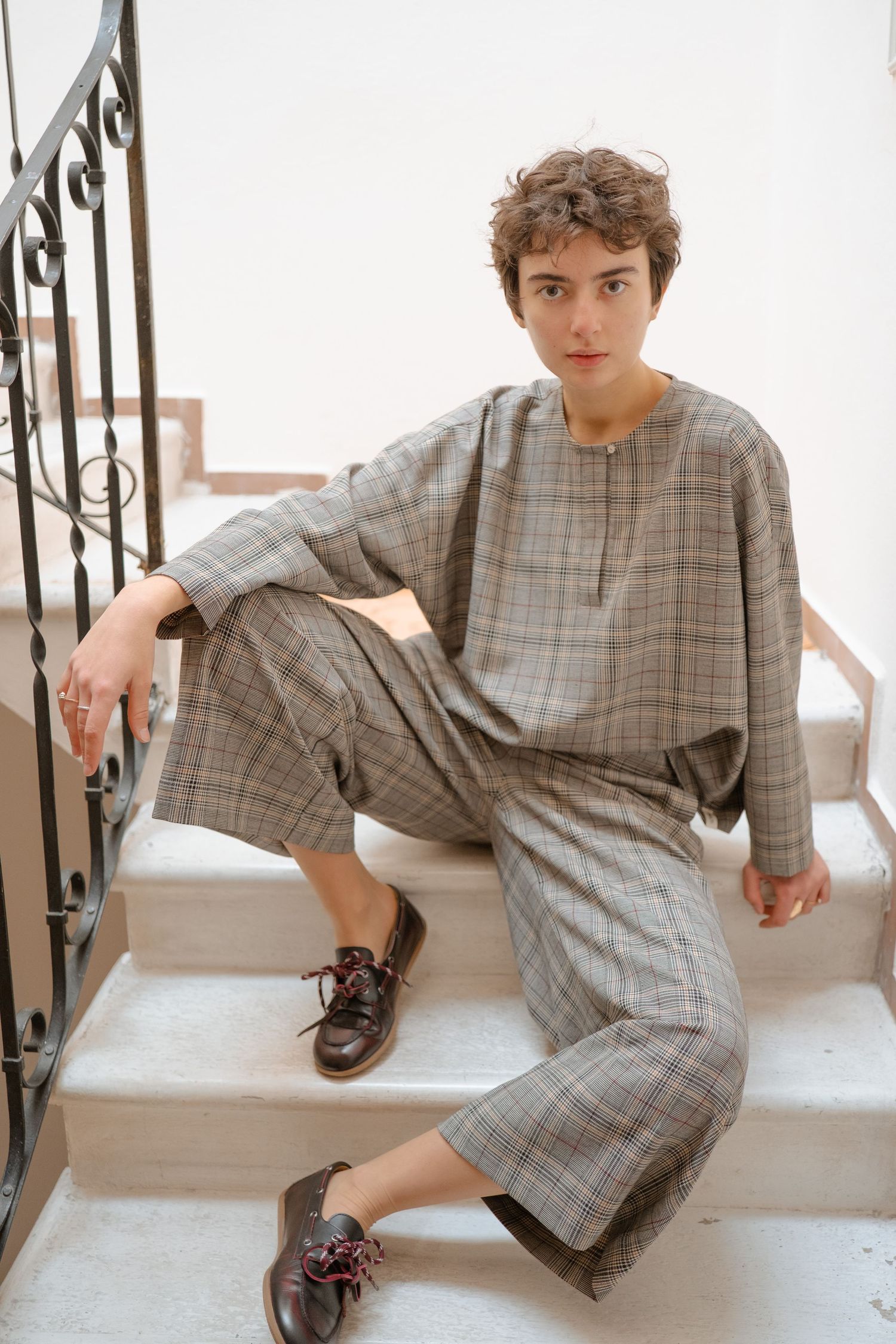 A person in a gray plaid jumpsuit lounges on white stairs in a series of casual poses.