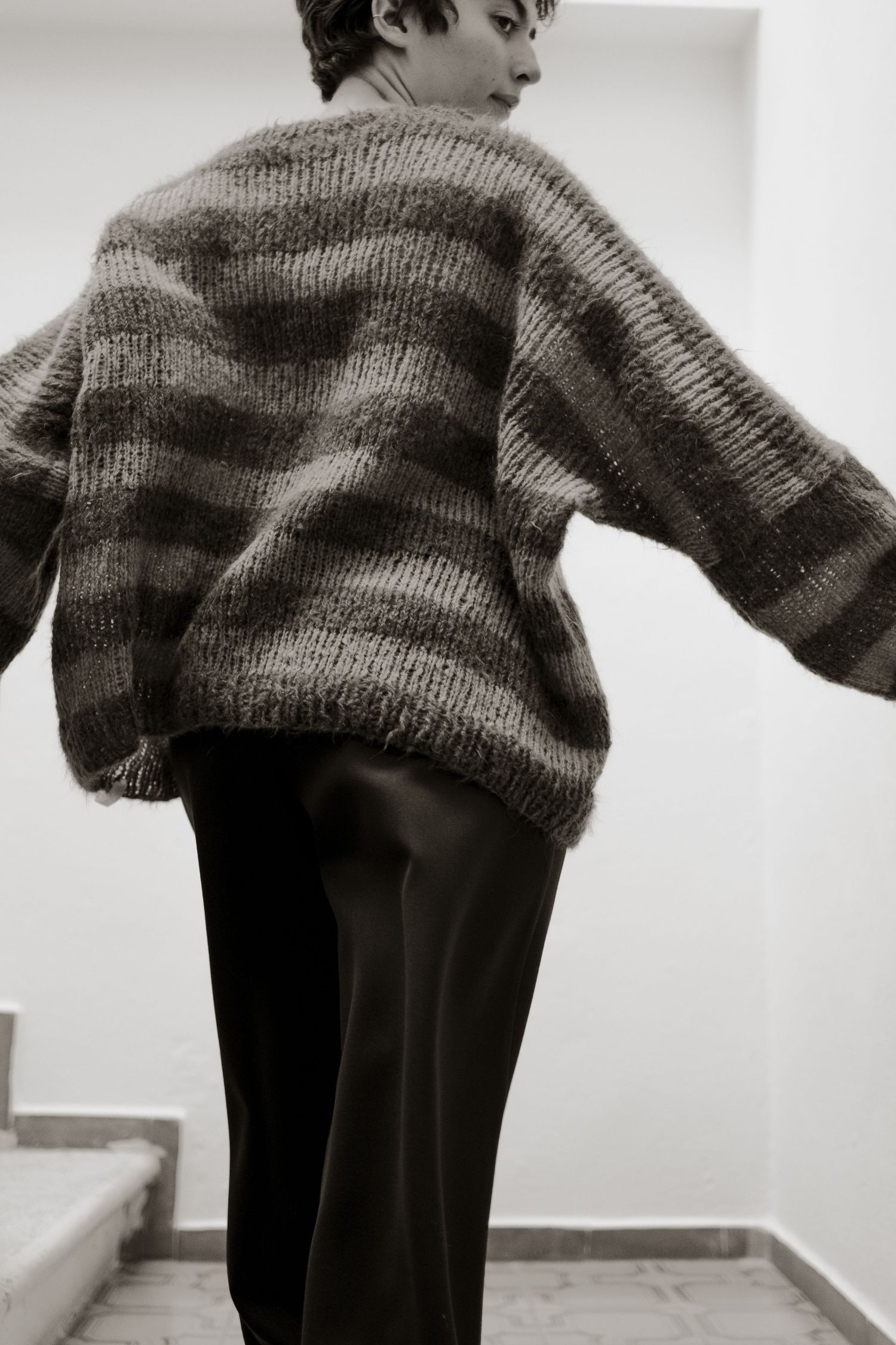 A person wearing a gray knit sweater and black pants stands against a white wall in a minimalist setting.