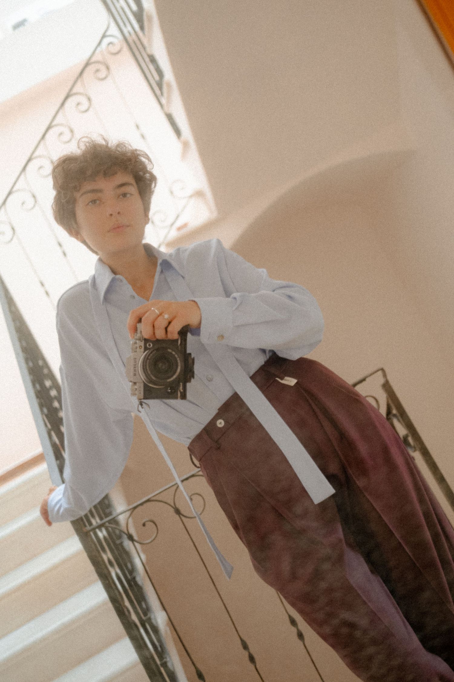 A person in a white button-down shirt and brown pants holds a vintage camera in a softly lit interior setting.