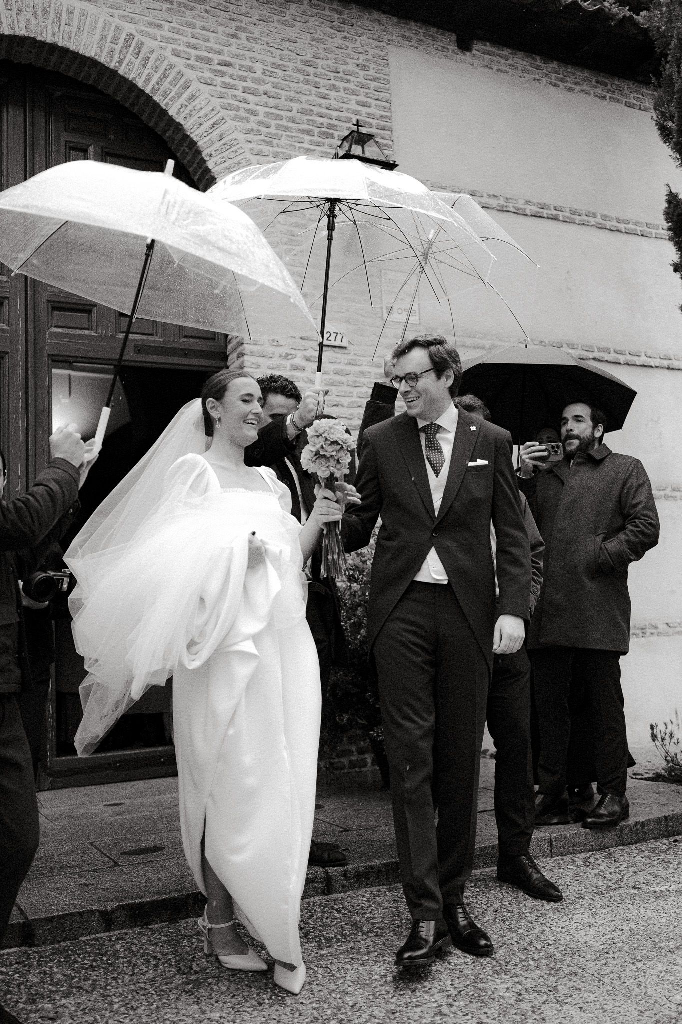 Wedding party walking under white umbrellas on a rainy day near brick building.