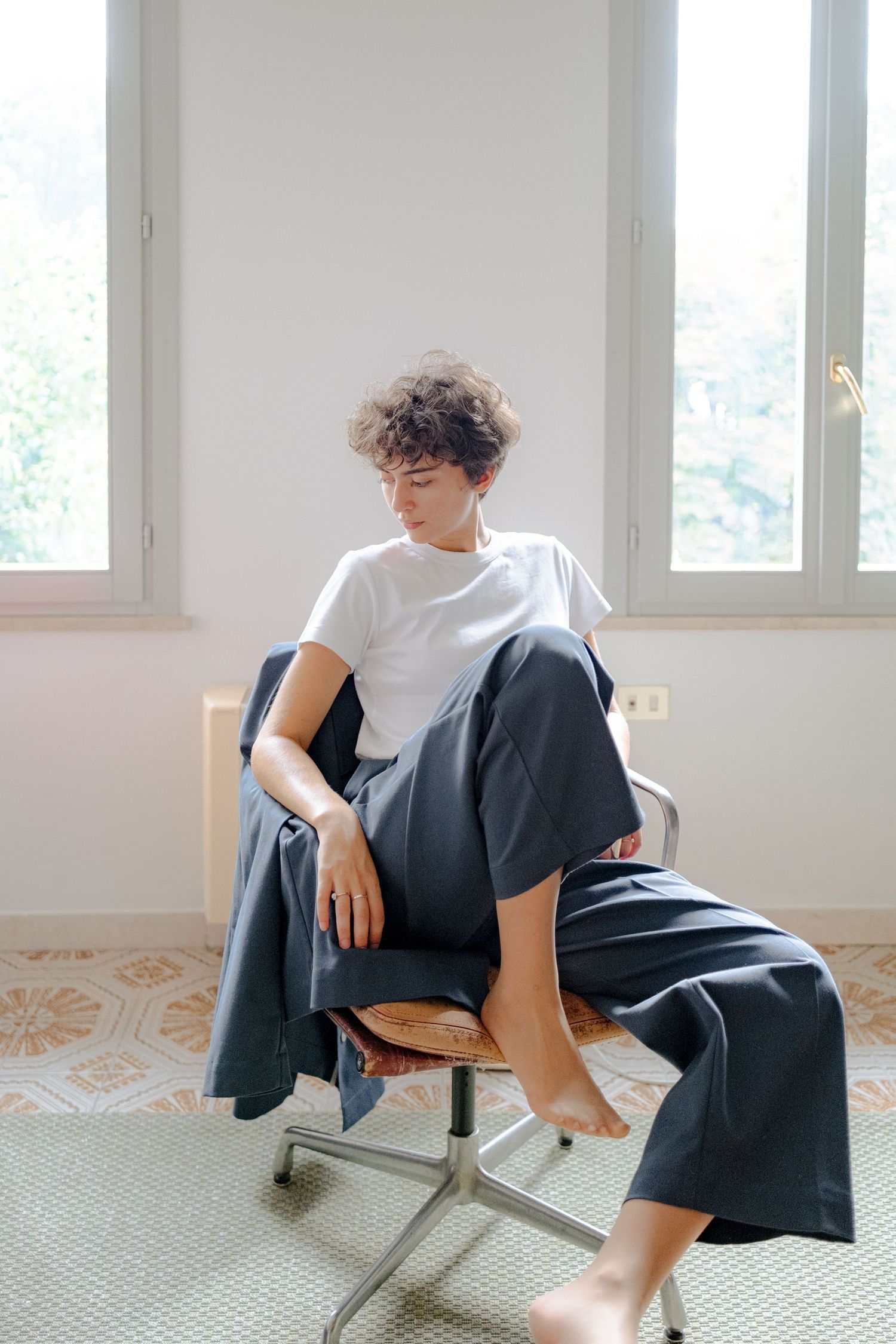 Person in white t-shirt and dark pants lounges casually on a modern chair against bright interior windows.