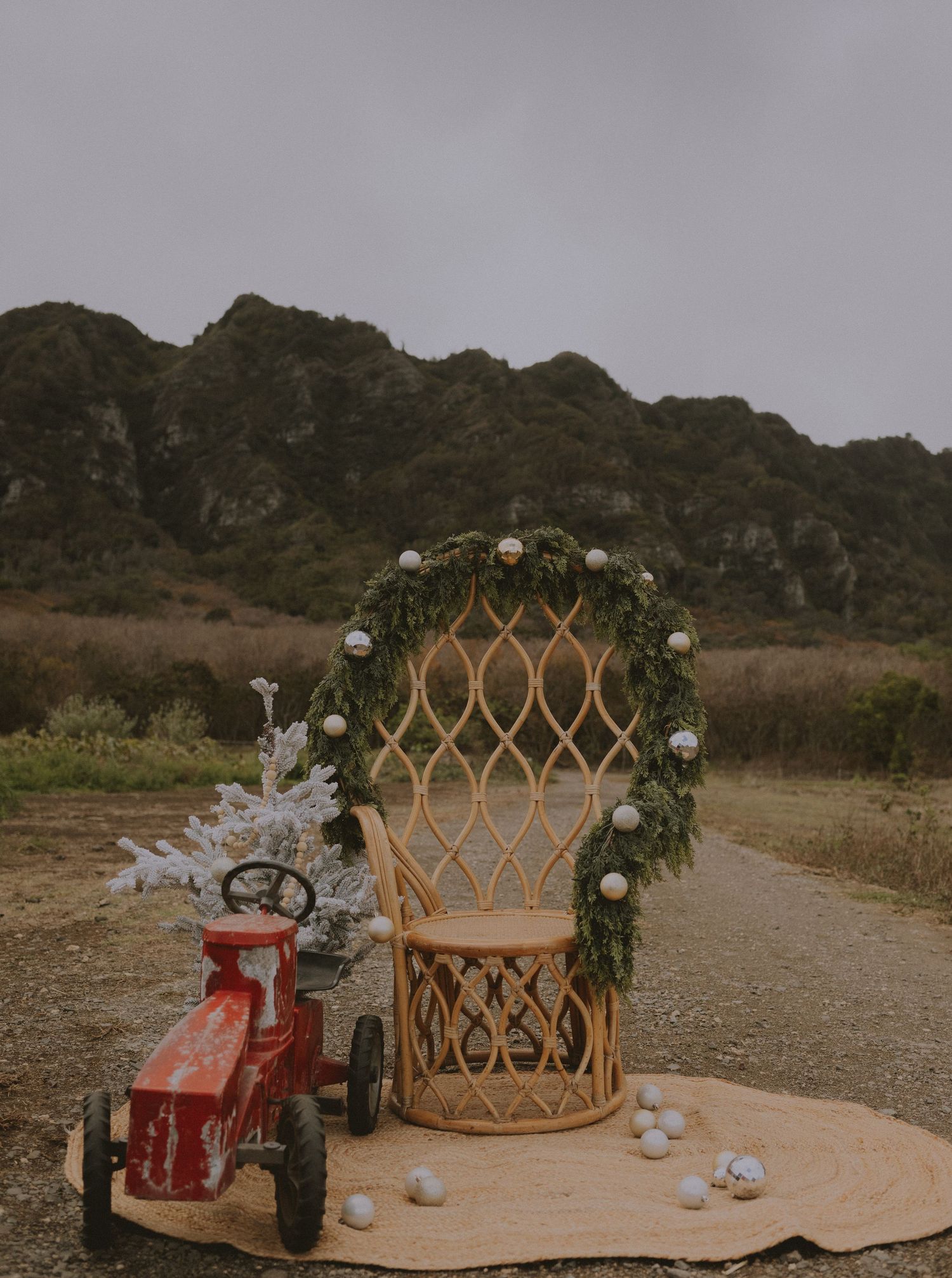 Decorated rattan peacock chair with garland arch and red vintage toy tractor on a dirt path.