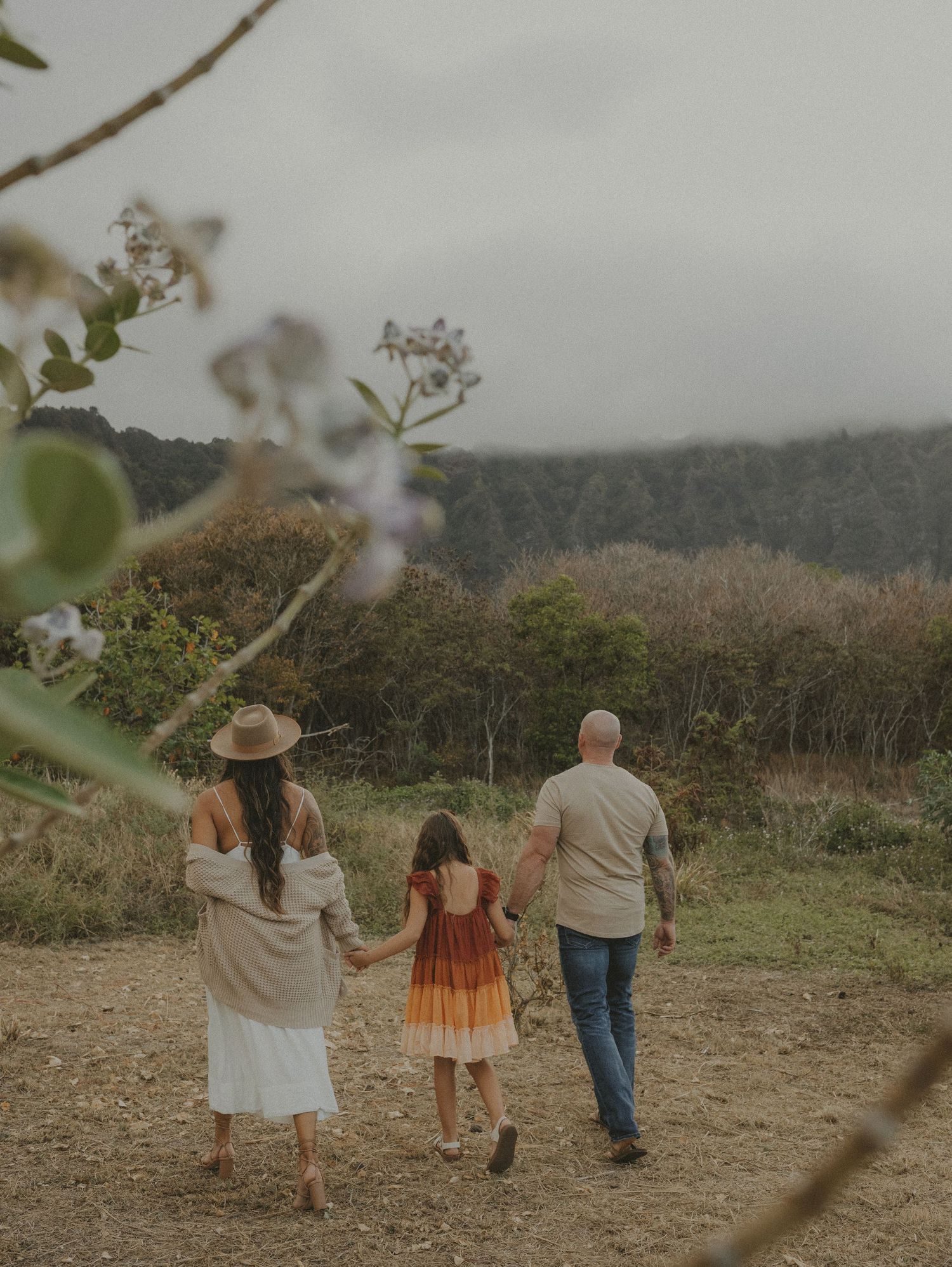 Family of three walking hand-in-hand through a scenic countryside trail with misty mountains ahead.