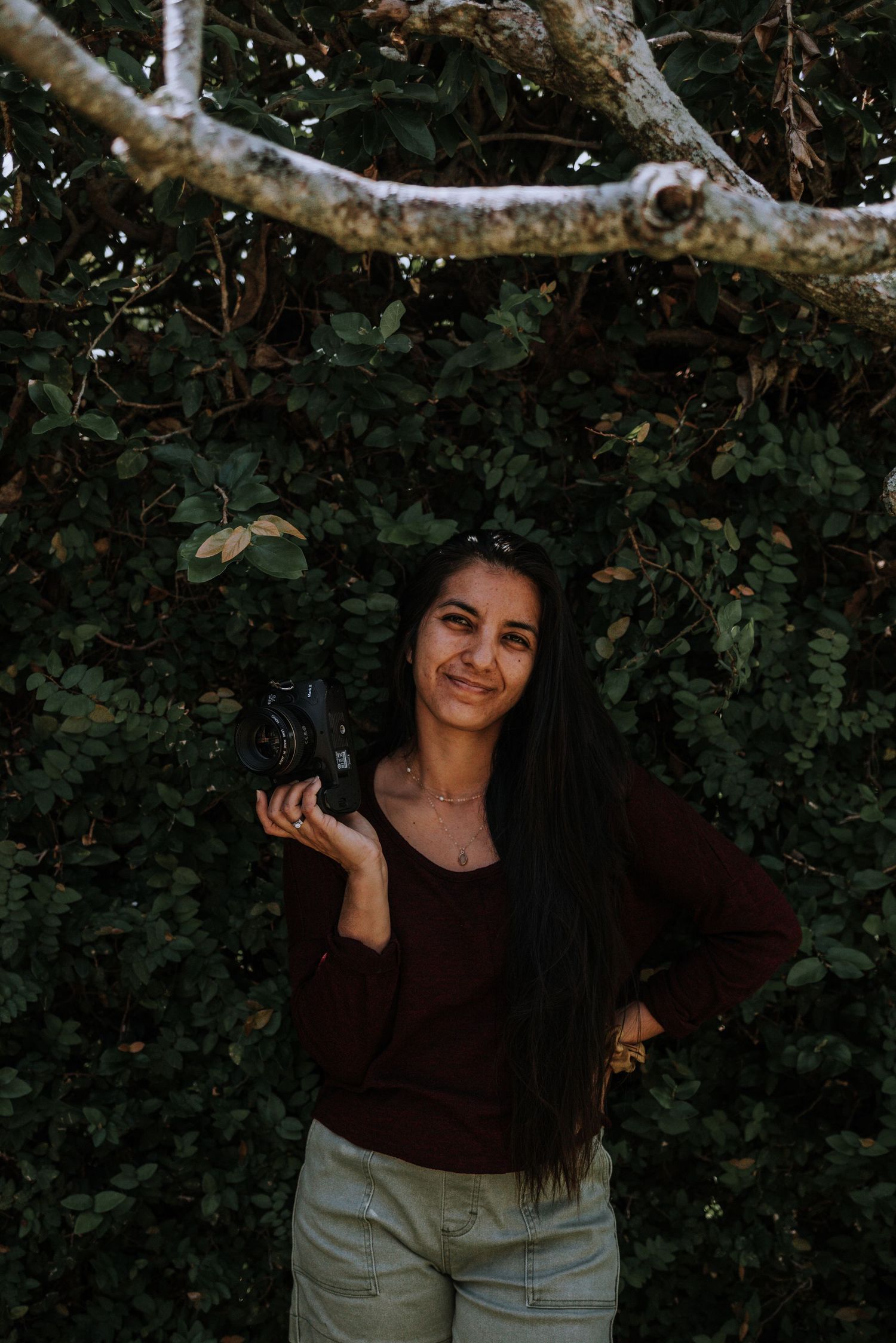 Smiling young female photographer holding a camera while standing in front of lush green foliage outdoors.