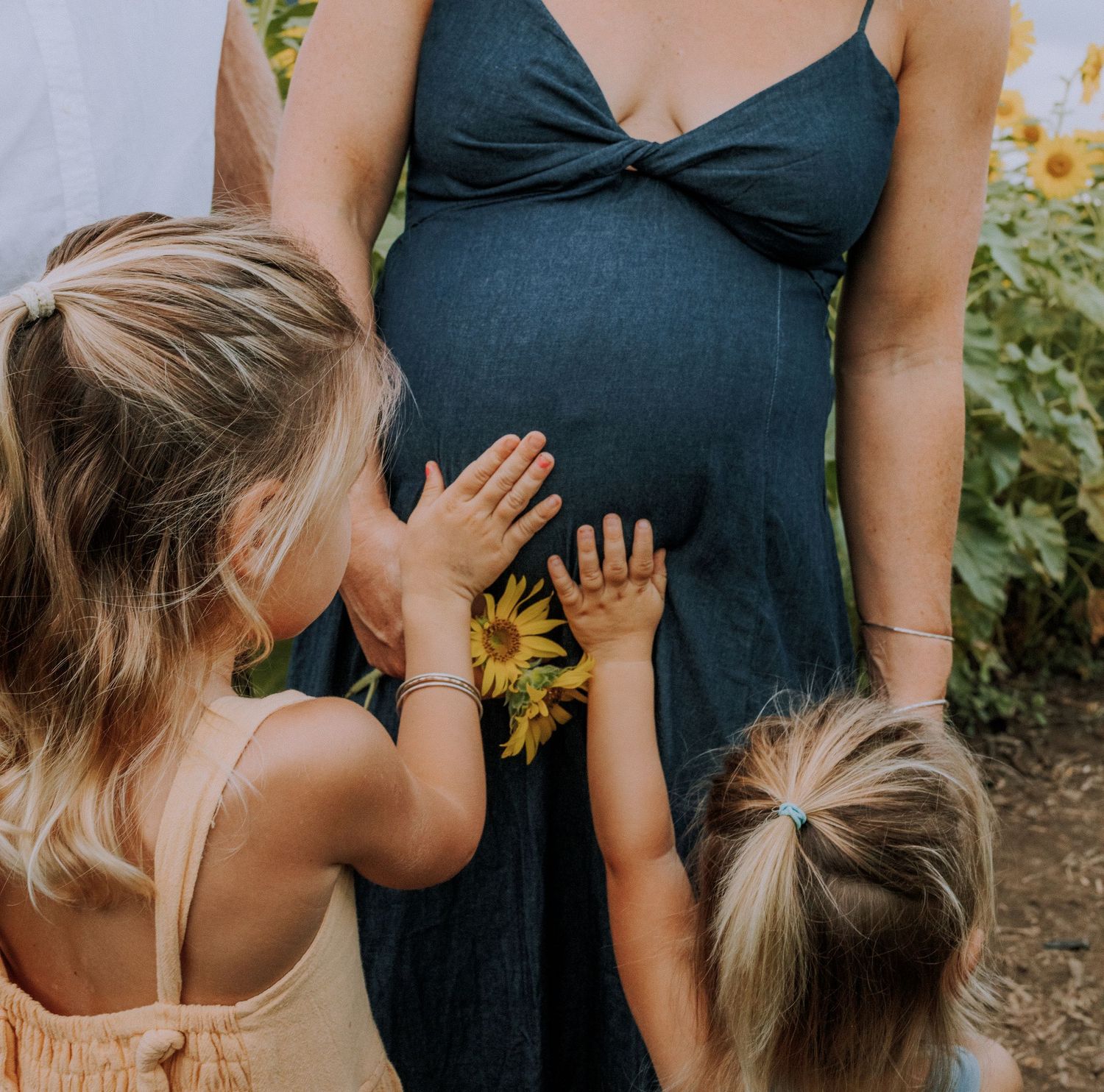 Pregnant woman in navy dress with two young girls touching her belly holding sunflowers in a sunflower field.
