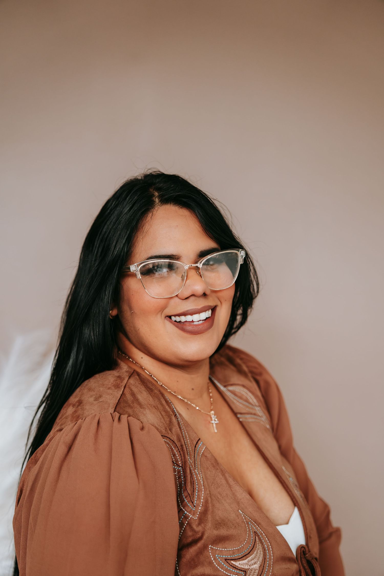 A smiling woman with long black hair and clear-framed glasses wears a brown embroidered blouse and gold necklace.