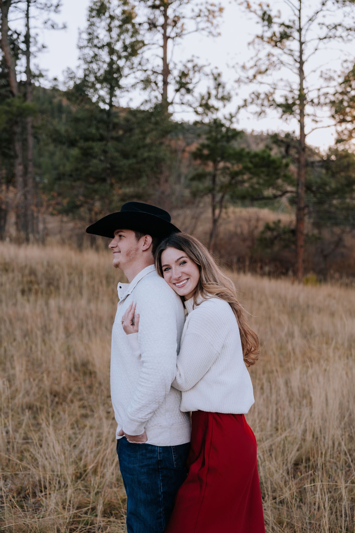 Woman in red skirt hugging man in cowboy hat from behind in a sunlit pine forest meadow.