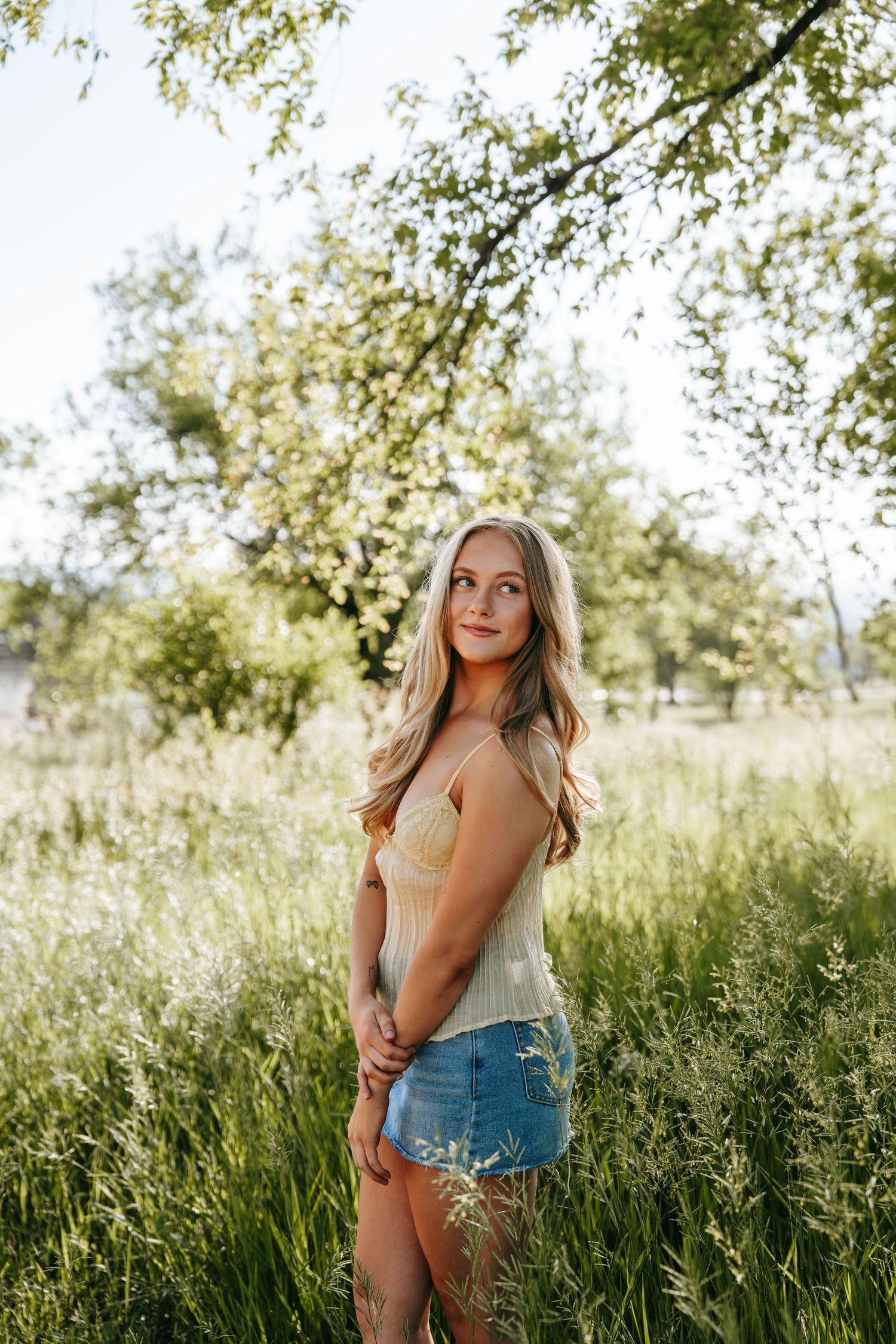 A young blonde woman in a yellow top and denim shorts stands in a sunlit green meadow beneath tree branches.