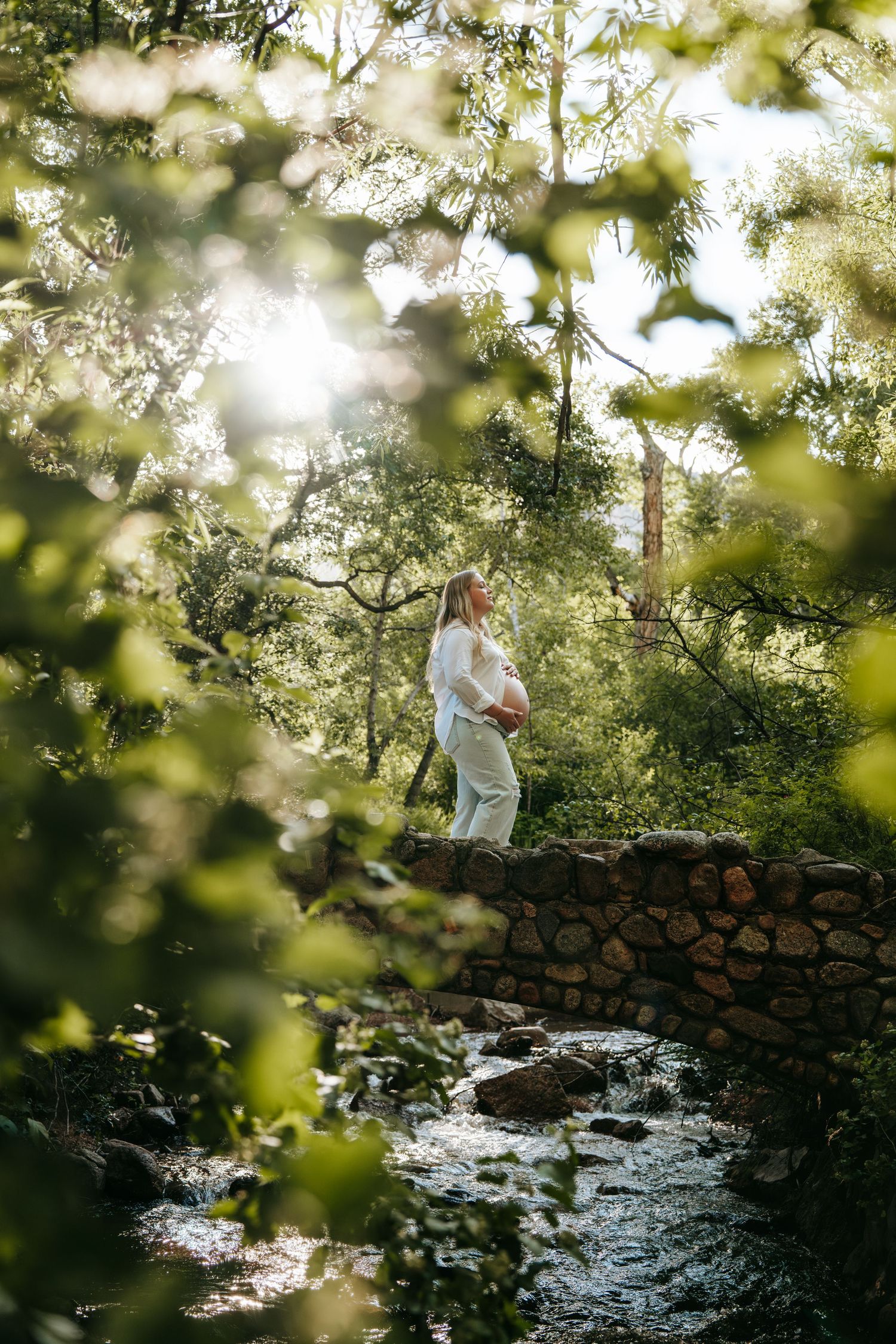 Pregnant woman stands on stone bridge surrounded by lush green forest in sunlight.