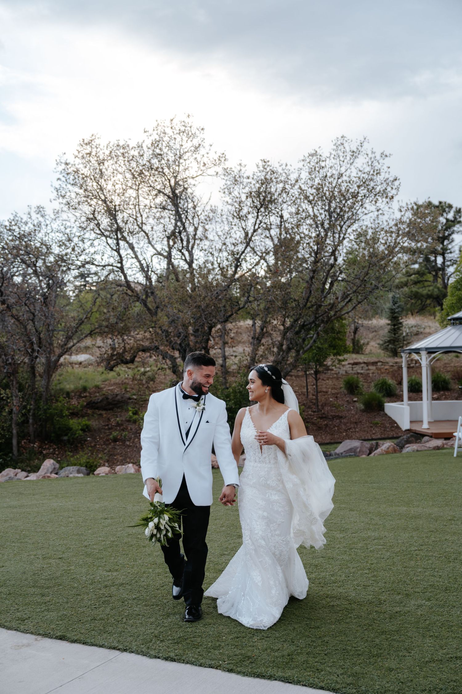Bride and groom walk hand in hand on green lawn, groom in white tuxedo, bride in fitted lace gown.