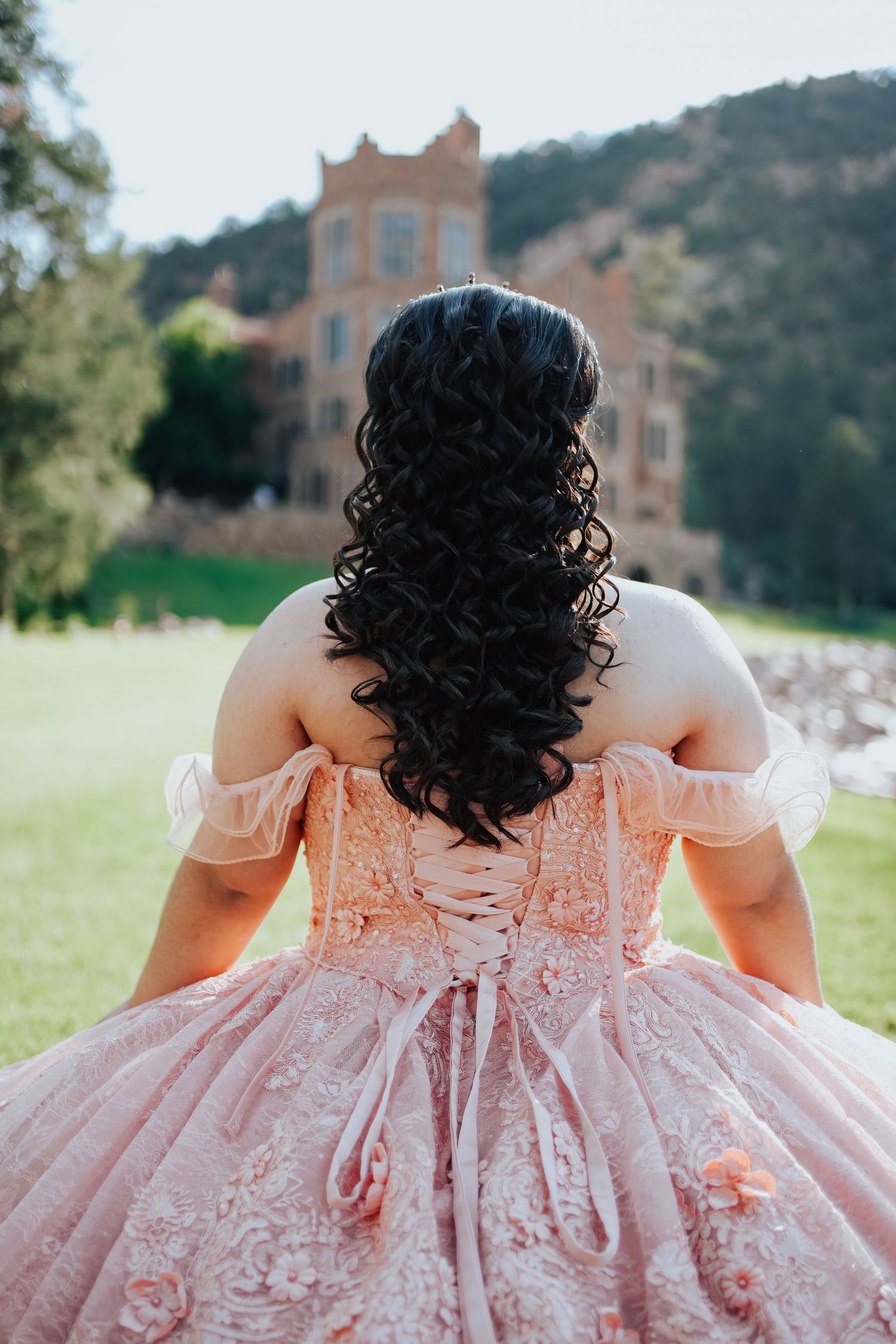 Back view of a young woman in a pink lace off-shoulder quinceañera gown before a castle.