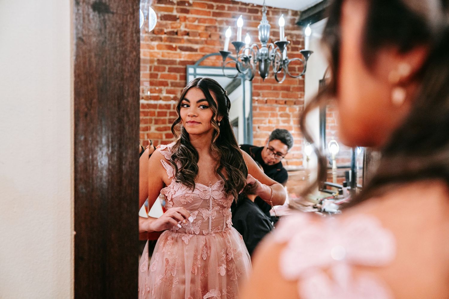 Young woman in pink quinceañera gown getting ready with hairstylist in rustic brick-walled venue.