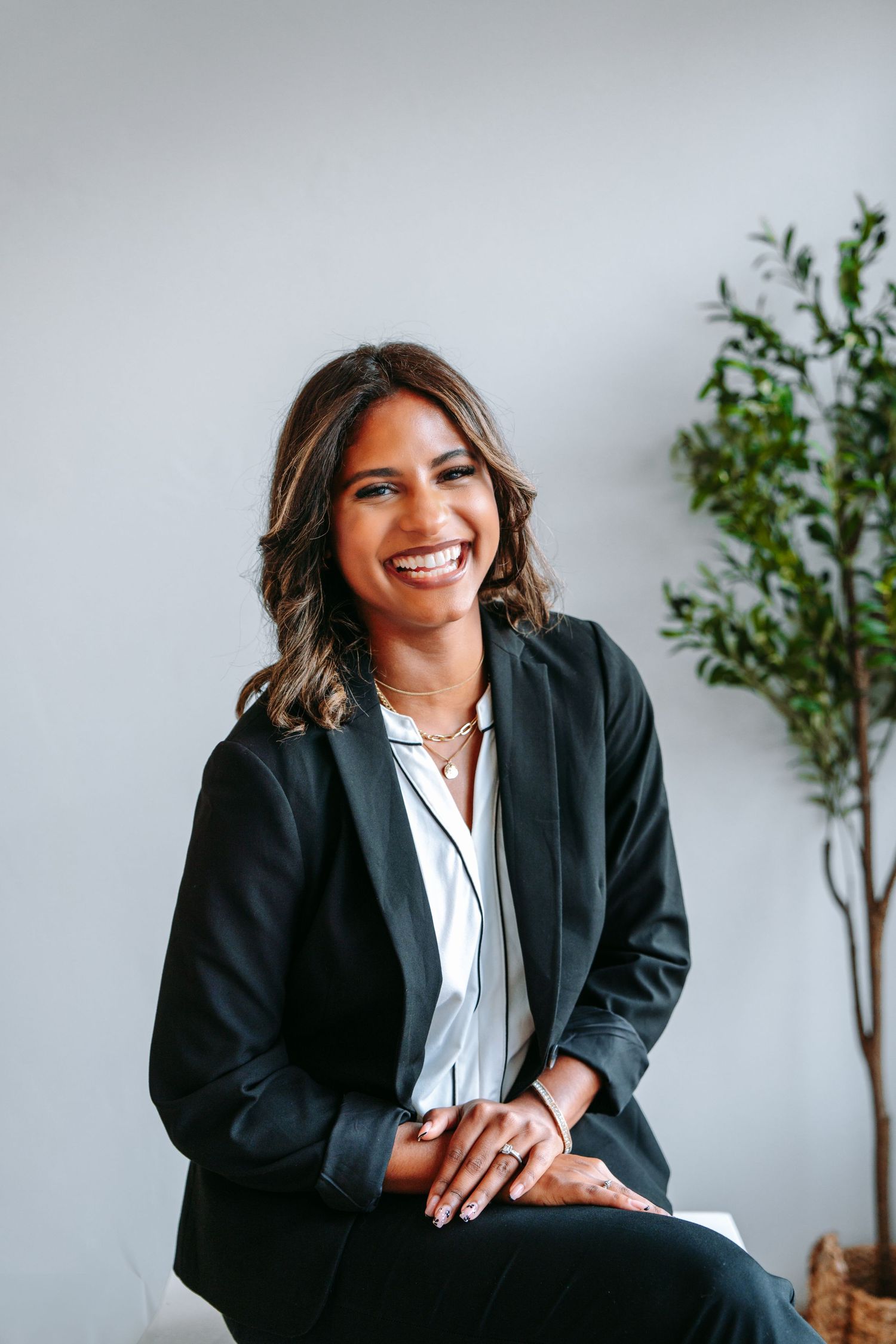 Cheerful professional woman in black blazer seated and smiling in a bright studio with a green plant nearby.