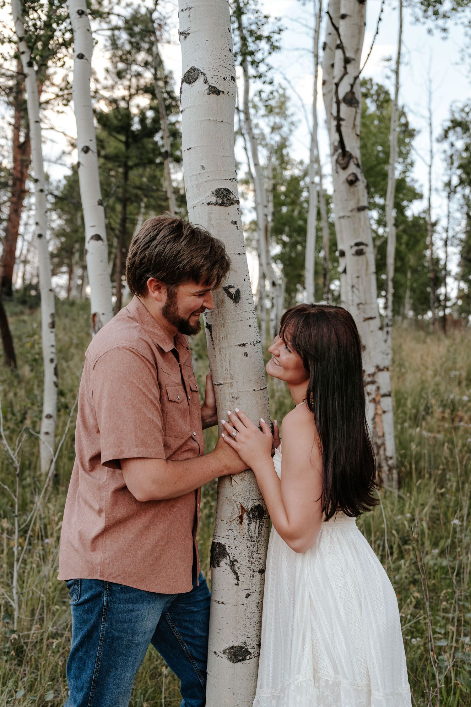Couple sharing a romantic moment among white birch trees in a lush forest, woman in white dress, man in pink shirt.