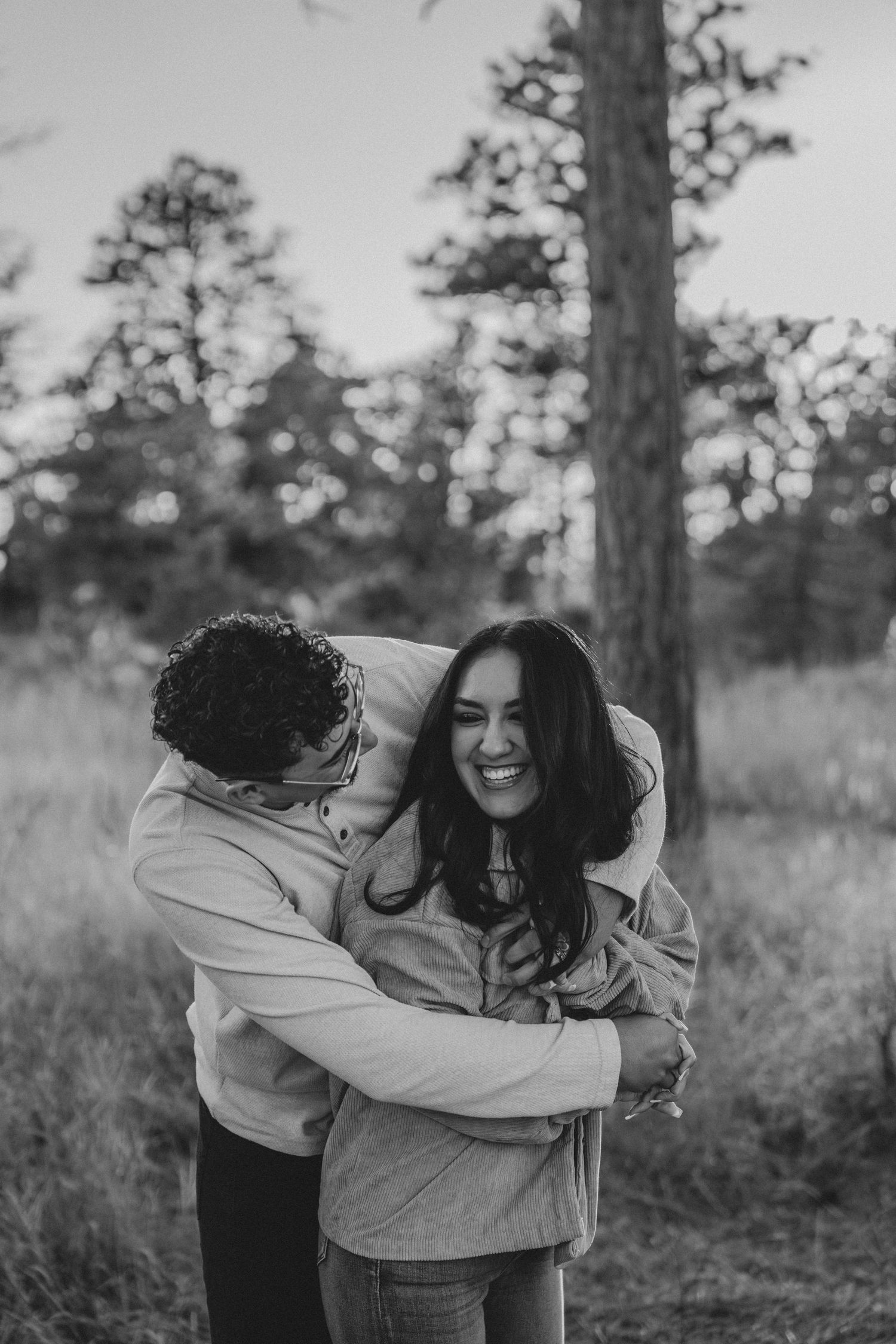 Black and white photo of a man embracing a laughing woman from behind in a pine forest setting.