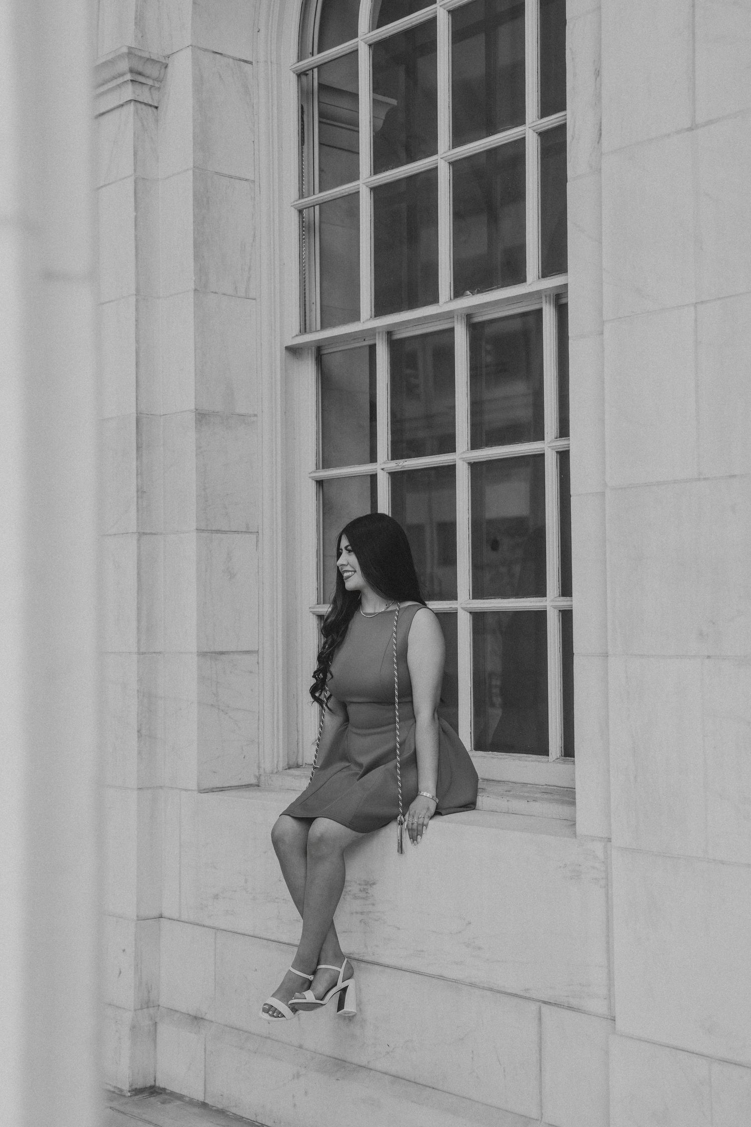 Black and white portrait of woman in dress leaning against marble wall beside large window.