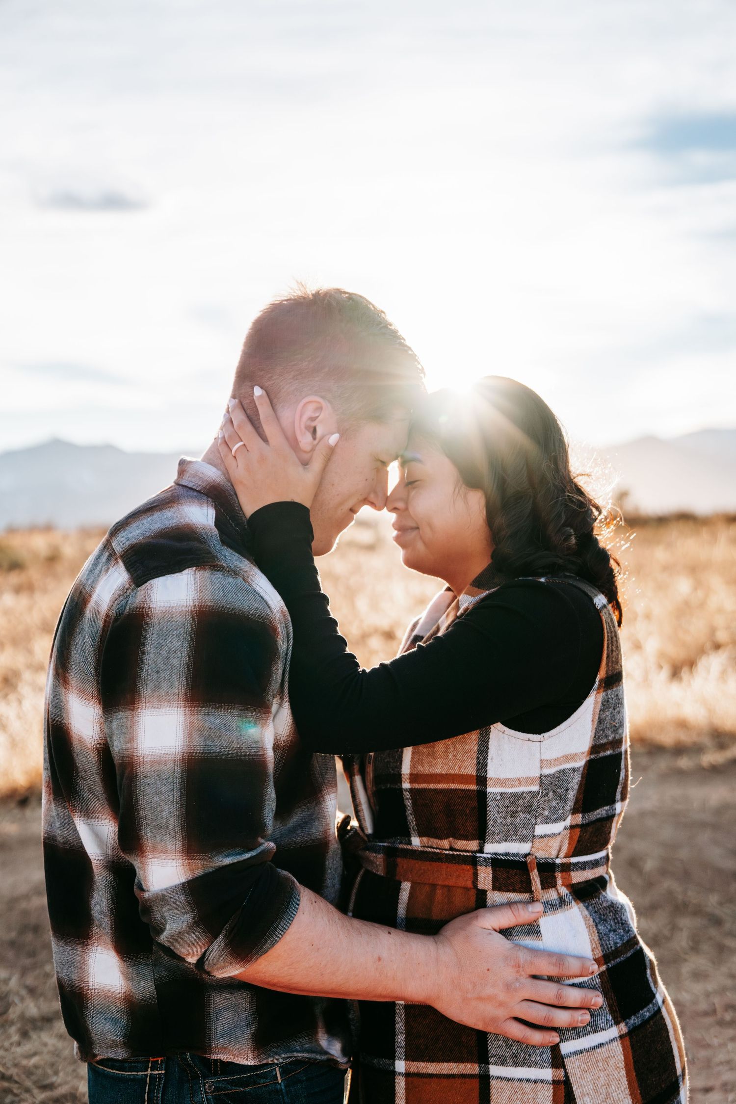 Pregnant couple touching foreheads romantically in a sunlit field with mountains behind them.