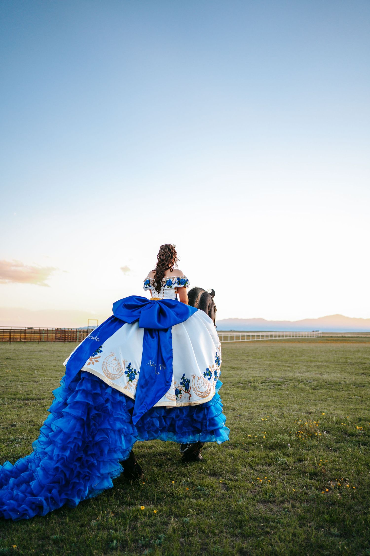 Quinceañera in blue ruffled ball gown rides black horse across green field at sunset.
